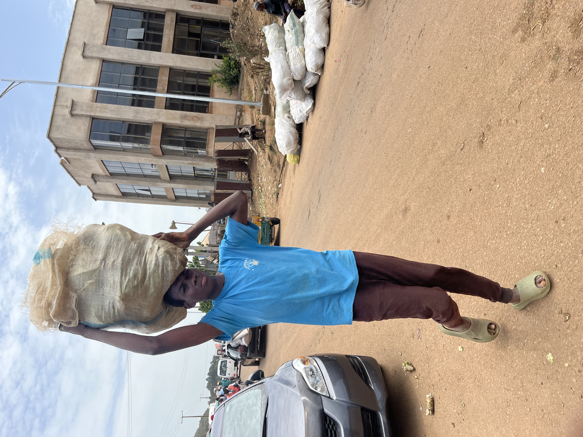 A young man carries a large burlap sack on his head. He is wearing a blue t-shirt and brown pants, with light green slides on his feet. The man is standing on a dirt and gravel road in front of a building with multiple stories and many windows. In the background, there are more burlap sacks piled up, some trees, and a few vehicles. The sky is cloudy, suggesting it may be daytime. The scene appears to be in a marketplace or a busy area where goods are being transported. The man's expression is neutral. There is no visible text in the image.