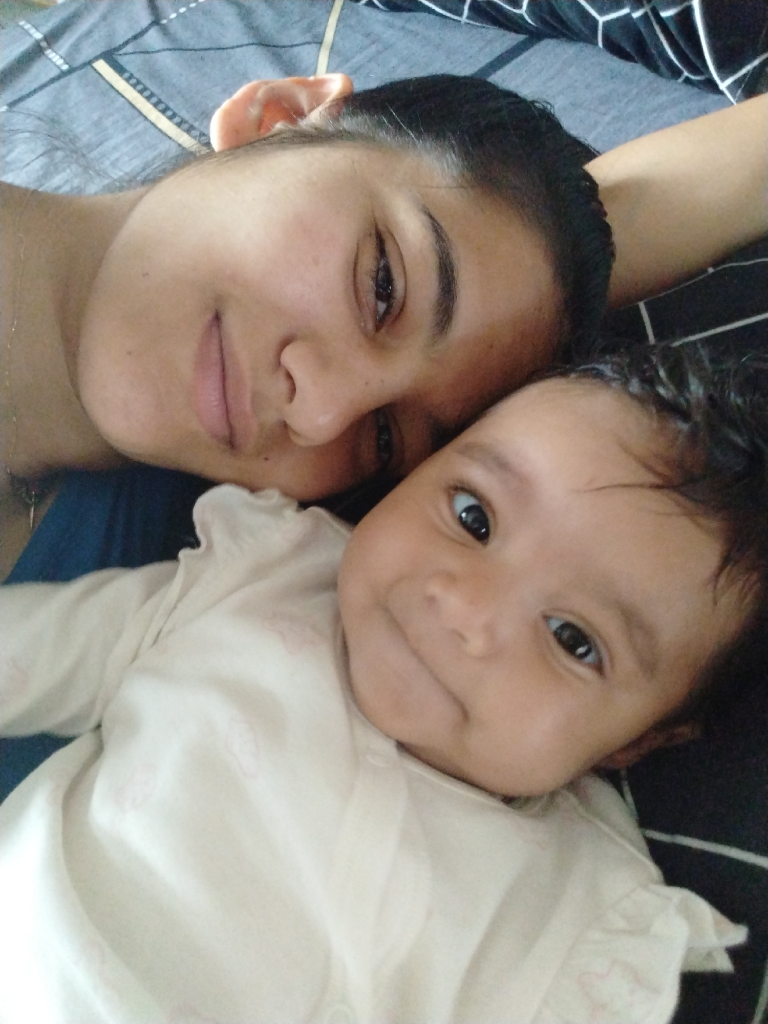 A selfie shot shows an adult female and a baby lying together on a bed with a black and white patterned sheet. The adult is on the left and is slightly out of focus. The baby, wearing a white shirt, is positioned on the right and is in sharper focus. Both the adult and the baby are looking at the camera with a slight smile. The city is Unknown, Unknown.