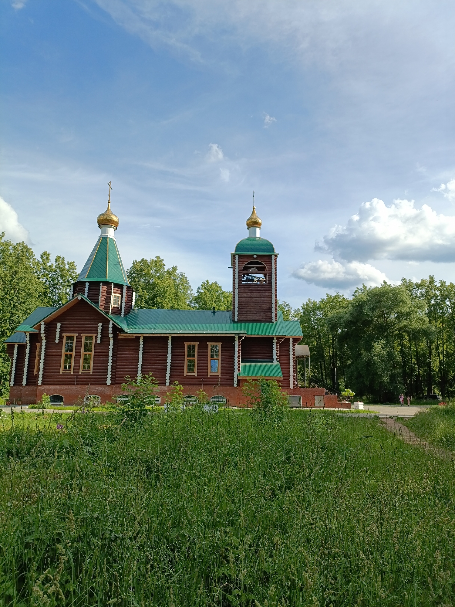 A wooden church with a green roof and golden domes stands amidst a lush green landscape. The church has a bell tower and is surrounded by trees. The sky is blue with scattered clouds, suggesting a pleasant day. In the foreground, tall grass grows abundantly, creating a natural and somewhat wild setting. In the background, a paved path leads away from the church, and a few people can be seen walking, indicating a public space or park. The overall scene evokes a sense of peace and tranquility. The architecture of the church suggests a Russian Orthodox style. The time of day appears to be daytime, possibly late morning or afternoon, given the brightness of the light and the appearance of the sky. The location is identified as Chaikovsky, Russia. There is no visible text in the image.