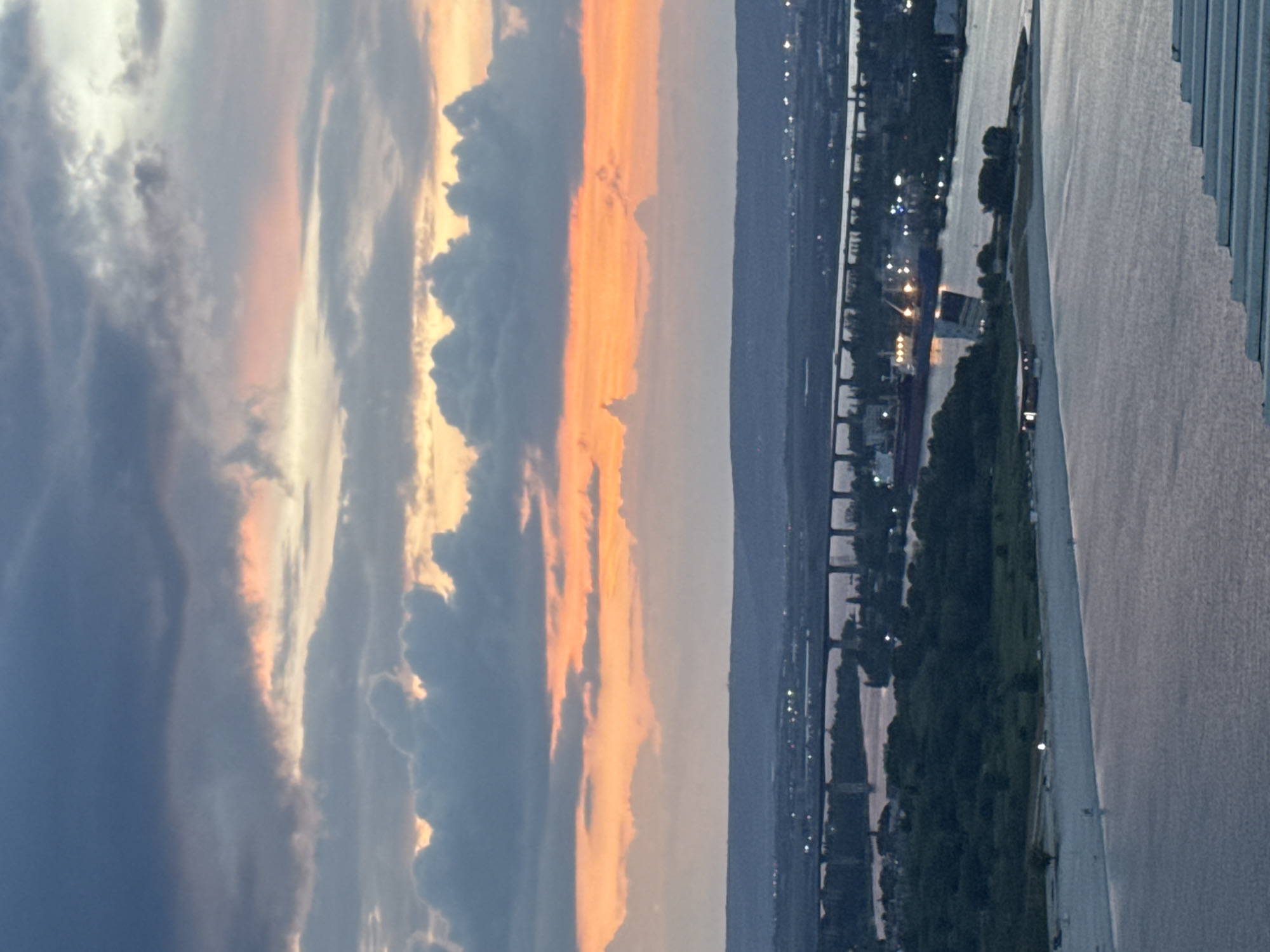 The image shows a sunset over the city of Varna, Bulgaria, seen from a high vantage point. The sky features layers of clouds colored with hues of orange, yellow, and dark blue. In the distance, a coastline is visible, with a line of city lights twinkling. Below, there are trees and a river or canal with bridges. The right side of the image features a body of water, possibly the Black Sea.