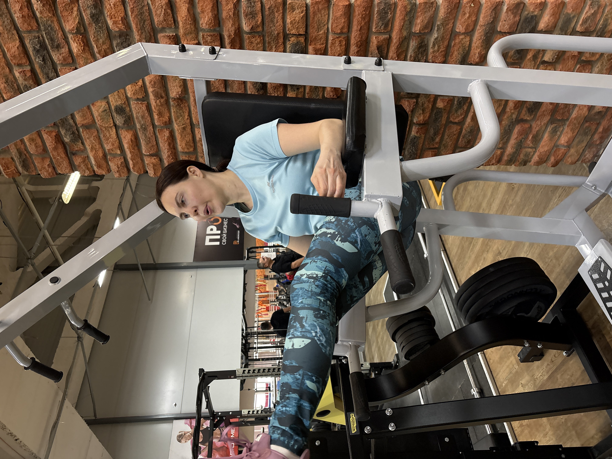 The image captures a woman using a weight machine in a gym. She is wearing a light blue t-shirt and patterned blue leggings. The weight machine is mostly gray and black, with black weights attached. The gym has a brick wall on one side and a commercial poster visible in the background. Other gym equipment and people are visible further in the background. The location is the city of Chaikovsky, Russia.