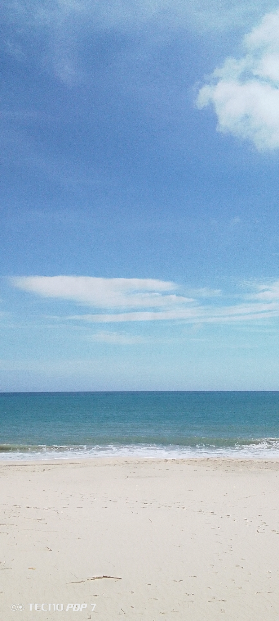 This is an eye-level, outdoor shot of a beach scene in Pedro González, Venezuela. The composition is divided into three main horizontal sections: sky, sea, and sand.
The sky occupies the top half of the image and is a clear gradient from a lighter, almost turquoise blue near the horizon to a deeper, more saturated blue at the top. There are a few wispy white clouds scattered across the upper part of the sky.
The sea is a band of turquoise-blue, meeting the sky at the horizon. Small white waves are visible as they approach the shore.
The beach stretches across the lower portion of the image, with light-colored sand. There are footprints visible on the sand. A thin, dark piece of driftwood is visible in the bottom-center.
The scene appears calm and peaceful. The image is taken under daylight conditions, with even lighting. There are no people visible in the shot. A "TECNO POP 7" watermark is visible in the lower left corner of the image.