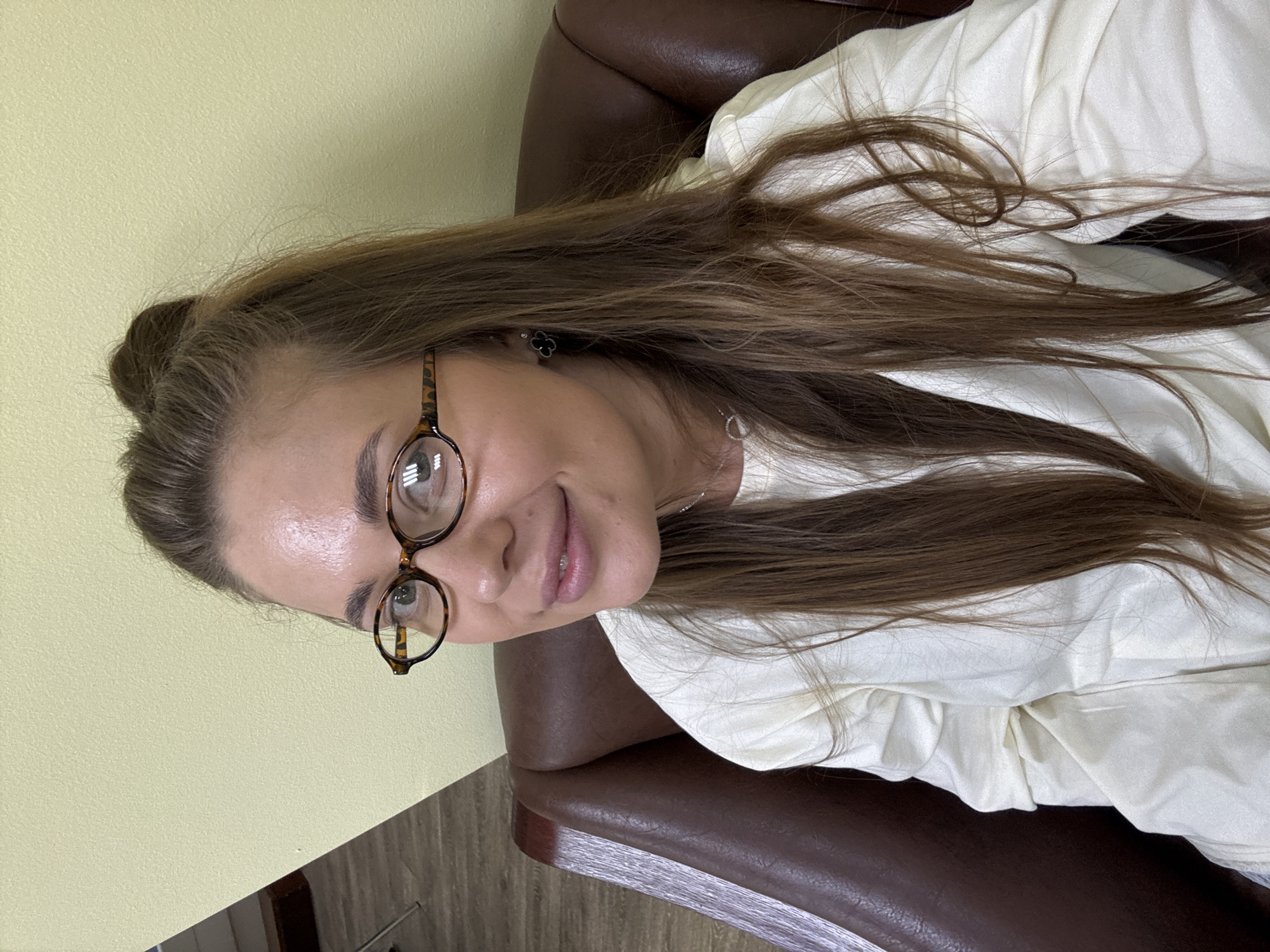 A woman with brown hair pulled into a bun is sitting in a brown leather chair. She wears tortoise-shell framed glasses and a light-colored shirt. The background consists of a light yellow wall. The location is Sosnovy, Russia.