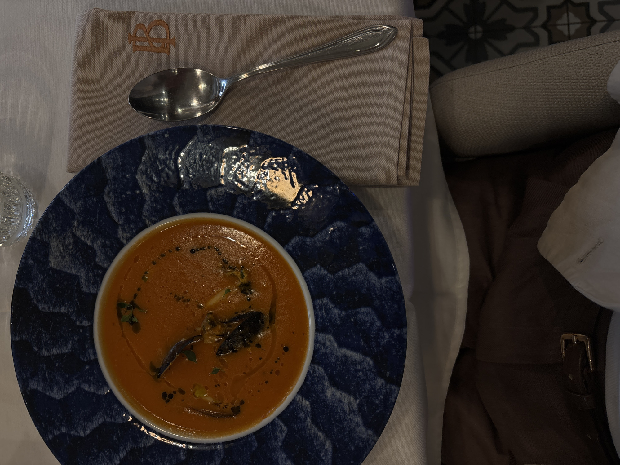 An overhead shot captures a dining scene in what appears to be a restaurant or upscale eatery in Yerevan, Armenia. The central focus is a bowl of rich, orange-red soup, possibly tomato or a bisque, garnished with green herbs, black specks (likely pepper or balsamic drizzle), and a few darker, elongated pieces, perhaps cooked vegetables or mushrooms, with a sheen of olive oil on the surface.
The soup is served in a white bowl, which rests on a dark blue, textured plate featuring a wave-like or abstract pattern. To the upper left of the plate, a silver spoon with an ornate handle lies on a neatly folded, light brown or beige cloth napkin. The napkin bears an embroidered monogram in a slightly darker shade, depicting two intertwined letters, clearly a capital 'B' and what appears to be a capital 'R'.
The table is covered with a crisp white tablecloth. A small portion of a clear, possibly crystal, drinking glass is visible on the far left. To the right of the dining setting, the background reveals elements of the environment. A section of floor or wall with dark grey and white geometric patterned tiles is visible. Below this, a beige upholstered chair or bench seat is present. Further to the right and bottom, dark brown fabric, possibly a jacket or bag with a gold buckle, and a white shirt sleeve or cuff are discernible, suggesting a diner is seated nearby. The lighting indicates an indoor setting.