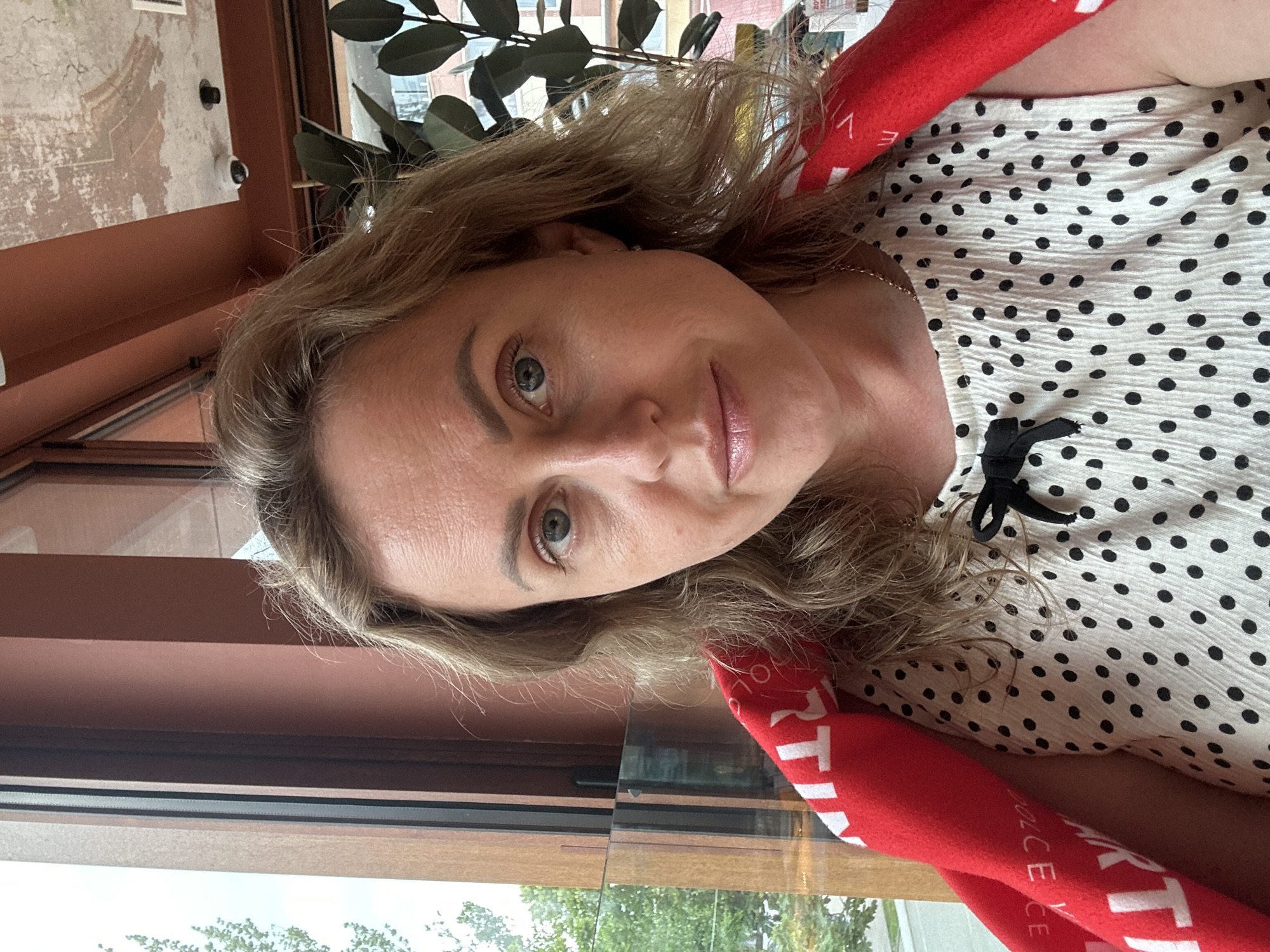 The image features a woman in a close-up, horizontally oriented selfie. She has blonde hair, light skin, and blue eyes, with makeup on her eyes and lips. She is wearing a white top with black polka dots and a black bow detail at the neckline. A red scarf or garment is draped around her shoulders, with visible white text that reads "ARTIKUL" and "DOLCE VITA". The background shows a window with a view of greenery and a hint of buildings, suggesting an indoor setting with an outdoor view, possibly a cafe or restaurant. The lighting indicates it could be daytime. The woman's expression is neutral, with a slight smile. There are no specific details indicating an event or activity other than the selfie itself. The location context is Anapa, Russia, which is not visually evident in the image itself.