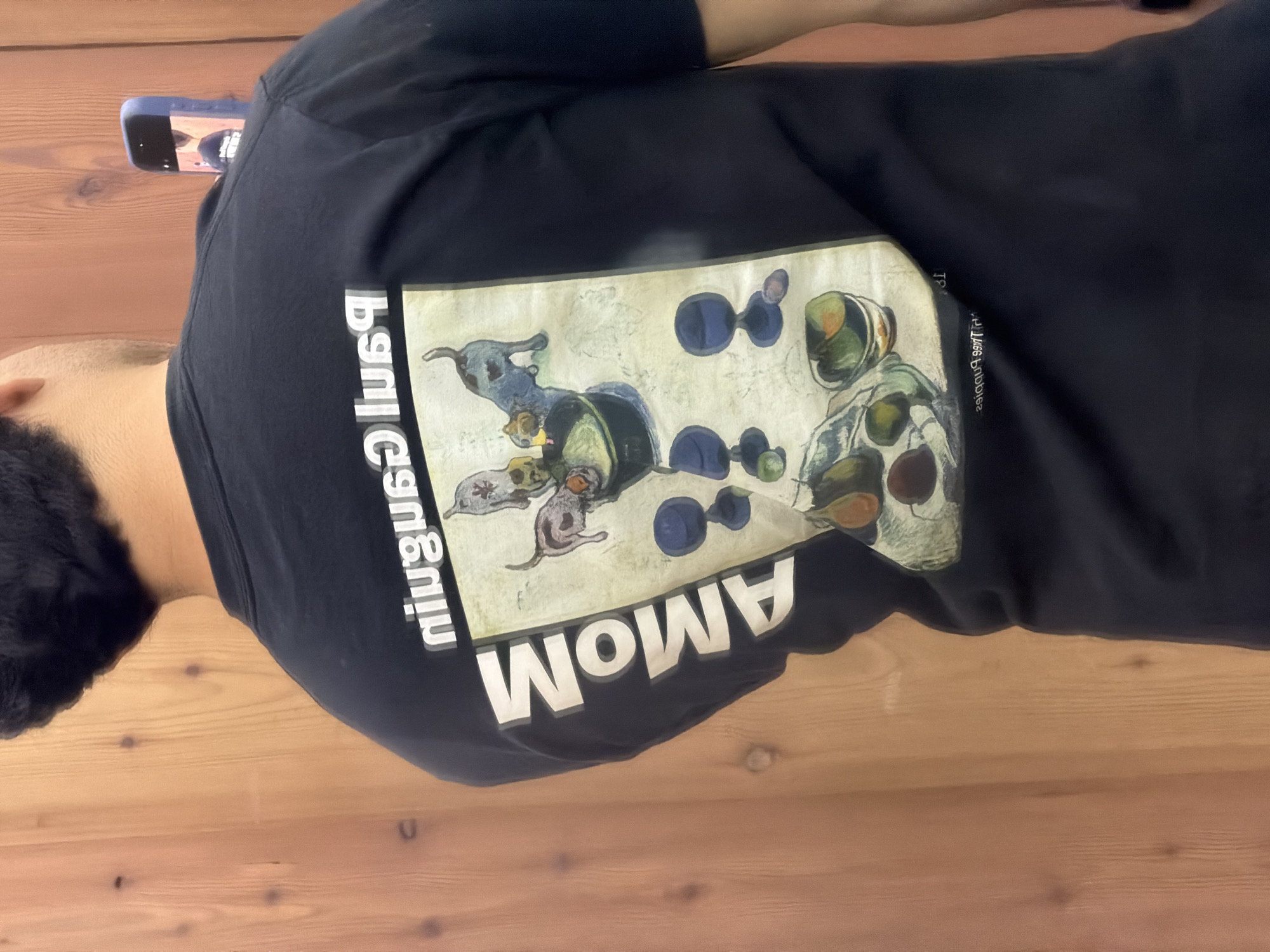 A person is wearing a black t-shirt with a graphic design. The design is off-white and features an illustration with dogs and circular shapes. Below the illustration, the word "AMOM" is printed in bold, white letters. The word "ninguga" is vertically printed in white beside the illustration. The photo is taken indoors on a wooden surface. A phone is visible at the top left of the frame.