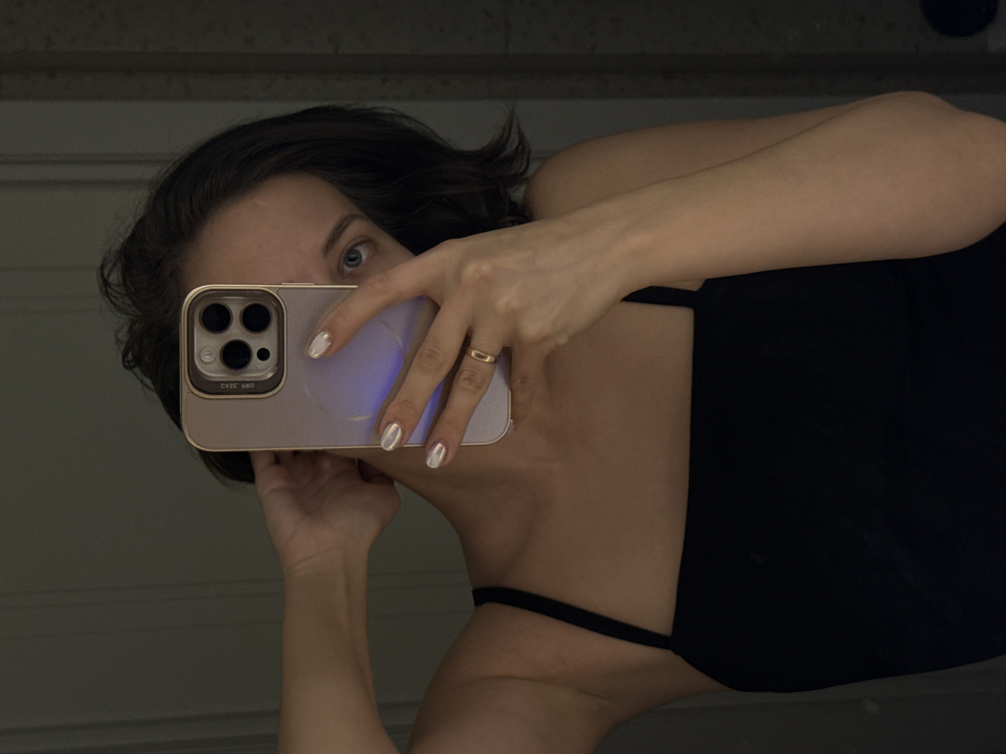 A mirror selfie captures a person, likely female, holding a smartphone to photograph their reflection. The individual has dark, short hair and a visible blue eye looking towards the camera. She is wearing a black top or dress with thin straps.
Her right hand holds a light-colored smartphone, possibly an iPhone, featuring a triple-camera system and a complementary light-toned case. Reflected purple light emanates from the phone's screen onto her fingers. She wears iridescent or metallic light-colored nail polish and a simple gold-colored band ring on her right ring finger. The phone case displays reversed text, including what appears to be "CYRE PRO" and "ОЯЗС ЭRО" (reversed "ORZASE" or similar).
The background behind the person is a dimly lit, plain wall, possibly with subtle horizontal paneling or texture, rendered in a dark grey or off-white tone. The overall lighting is low, indicating an indoor setting, potentially at night or in a room with subdued illumination. The image provides a casual, self-portrait perspective. Based on external information, the location context for this image is Pervomaysky, Russia.