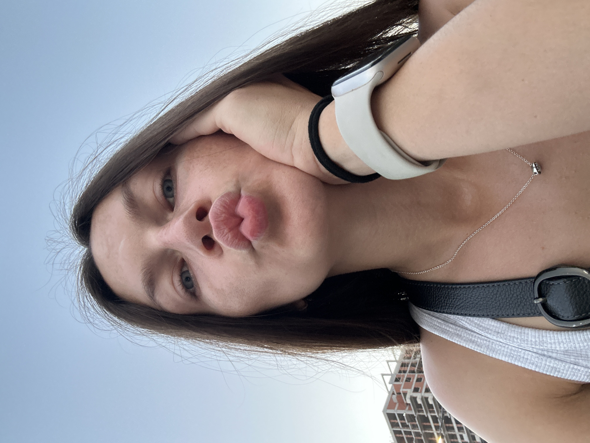 The image is a close-up, selfie-style photograph of a young woman with long, dark brown hair, looking directly at the camera with her lips pursed in a kiss gesture. She is outdoors, with a clear sky in the background. A partially constructed building, possibly a hotel or apartment complex, is visible on the left side of the frame, suggesting an urban or developing area. The woman is wearing a light-colored tank top and a silver necklace. A black strap, likely from a bag, crosses her chest, and she is wearing a smartwatch on her left wrist. Her right hand is resting on her cheek, near her eye. The lighting suggests it is daytime, possibly late afternoon or early evening due to the soft light. The location is identified as Budva, Montenegro.