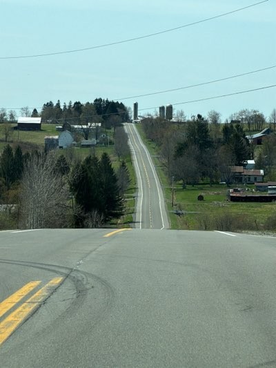 The image presents a daytime view of a rural, two-lane asphalt road stretching into the distance through a hilly landscape, likely in Groton, United States. The road, marked with double yellow lines in the center and white lines on the shoulders, appears to dip immediately in front of the viewer before ascending steeply over a distant hill. Tire marks are visible on the asphalt in the immediate foreground, to the left of the yellow lines.
On both sides of the road, the scene is characterized by scattered farmsteads and clusters of trees. To the left, a large barn with a dark roof and a lighter-colored house are visible, surrounded by mostly bare deciduous trees and some evergreens. Further up the road on the left, more structures are discernible. On the right side, multiple farm buildings, including houses and several prominent tall, cylindrical silos, stand out against the clear blue sky near the top of the distant hill. A large round hay bale can also be seen in a grassy field on the right.
Overhead, multiple utility lines traverse the sky, stretching across the frame. The trees, many of which are bare, suggest the season is either early spring or late fall. The sky is bright and clear, indicating fair weather. No people are visible, and there is no discernible text within the image. The scene conveys a sense of peaceful, open countryside.