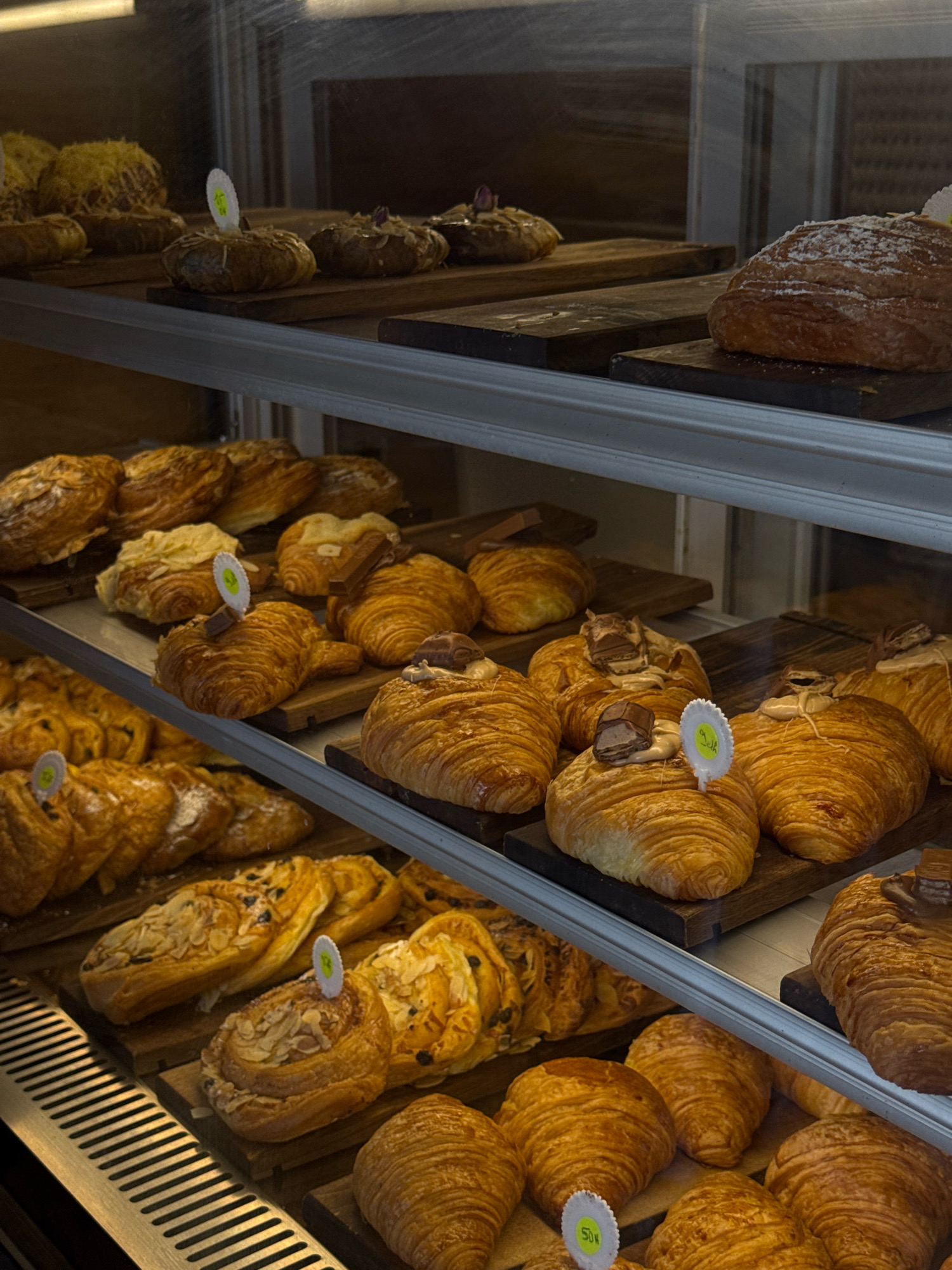 The image displays a refrigerated glass display case filled with a variety of pastries. On the top shelf, there are several round pastries topped with shredded cheese and nuts, as well as a large croissant dusted with powdered sugar. The middle shelf showcases an assortment of croissants, some plain, others with what appears to be chocolate or cream filling, and one topped with a piece of chocolate. The bottom shelf is laden with more croissants, including some sprinkled with almonds and raisins, and a few that are plain. A metal grate runs along the bottom of the display case. The lighting is dim, casting a warm glow on the baked goods. The scene is likely a bakery or pastry shop in Essaouira, Morocco, given the context provided.