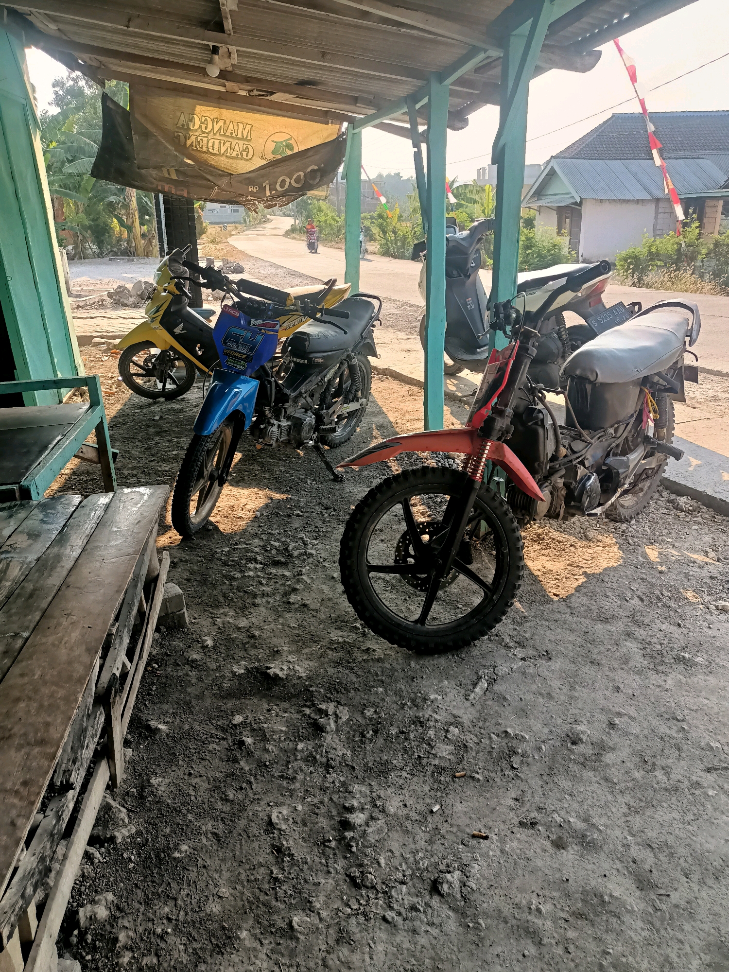 The image depicts an outdoor scene in Sumber Bendo, Indonesia, likely a roadside stall or a simple garage. In the foreground, a weathered wooden bench is visible on the left. Several motorcycles are parked under a covered area with a corrugated metal roof supported by green-painted wooden posts. The ground is a mixture of dirt and gravel, with some debris.
The motorcycles vary in color and style. One is bright yellow, another is blue, and a third is predominantly red with black accents and knobby tires, suggesting it may be an off-road or modified bike. The blue motorcycle has graphics and text on its front panel. The red motorcycle has a license plate that reads "S 5235 AAU". In the background, a road curves away, with a person riding a motorcycle in the distance. Houses with red and white flags are visible on the right side of the image, indicating a possible celebration or national holiday. A banner with text and a logo hangs from the roof of the covered area, further suggesting a commercial establishment. The lighting indicates it is daytime, possibly late afternoon, with sunlight casting shadows.