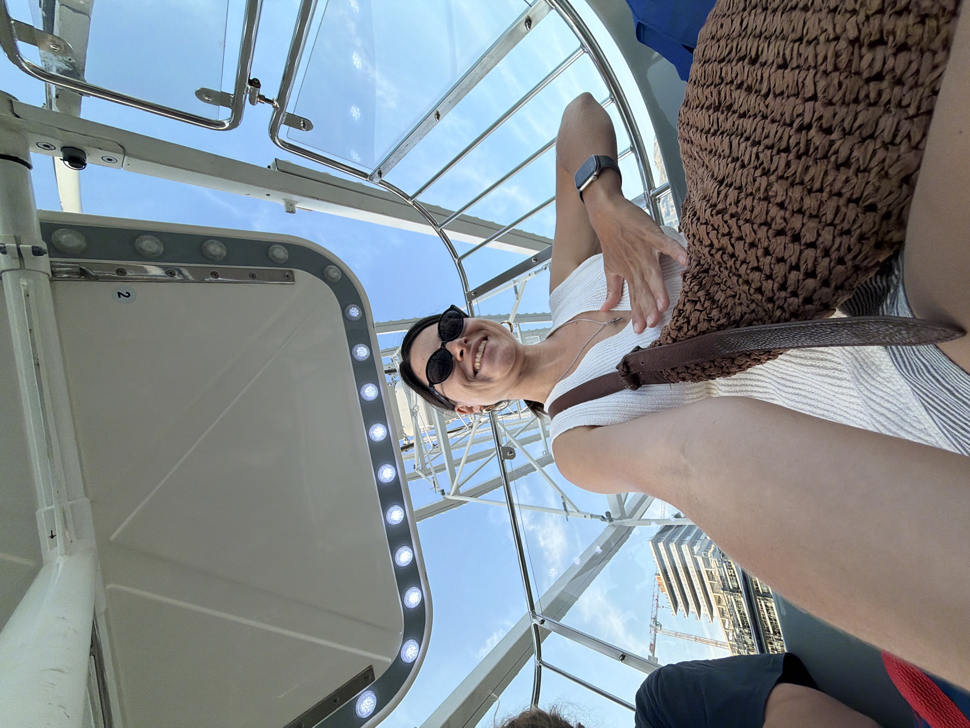 The image captures a low-angle perspective inside a modern, transparent observation capsule, likely part of a Ferris wheel, during daytime. The primary subject is a smiling woman, looking directly at the camera. She has dark hair, is wearing sunglasses, a white ribbed top, a silver necklace with a pendant, and a smartwatch on her left wrist. A brown, textured bag with a strap rests on her lap or beside her. Part of another person's leg or knee is visible in the bottom right corner.
The interior of the gondola is white with sleek design. A row of circular LED lights illuminates an inner panel, and a metal handrail is also visible. Through the clear ceiling and walls, a bright blue sky with scattered clouds is prominent, indicating sunny weather. Outside the capsule, the white metal structure of the ride itself is visible. In the background, an urban environment features a multi-story building under construction, complete with scaffolding and cranes.
A small white label displaying the number "2" is affixed to the interior wall of the gondola. The woman's joyful expression suggests she is enjoying the ride and the views from this elevated vantage point in Budva, Montenegro.