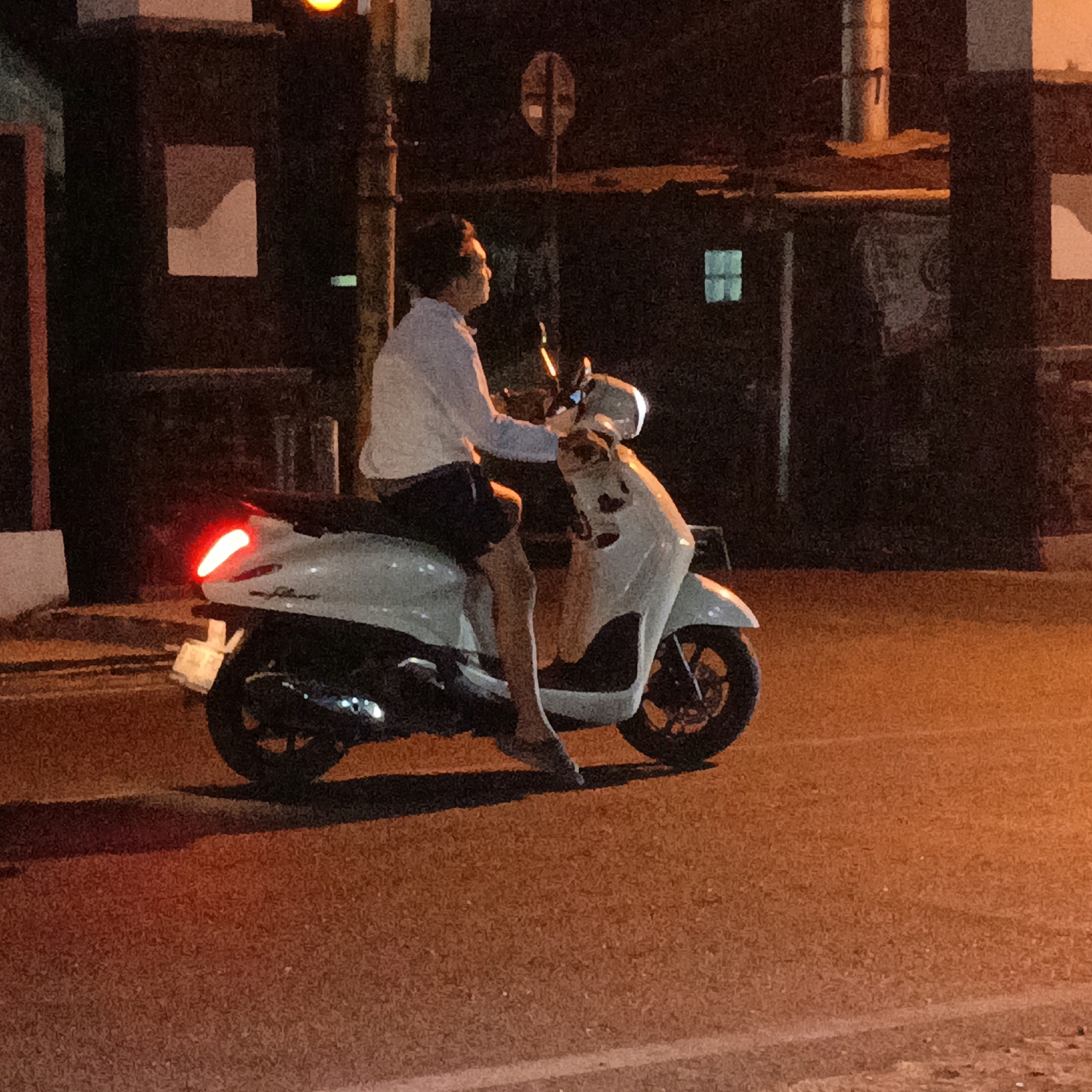 A person rides a white scooter at night on a city street in Salatiga, Indonesia. The scooter has a red taillight and a license plate. The rider is wearing a light blue shirt and dark shorts, with their right leg extended. They are looking to their left. The street is paved and appears wet. In the background, there are dark buildings with some illuminated windows and a traffic light that is yellow. The overall lighting suggests it is after dark.