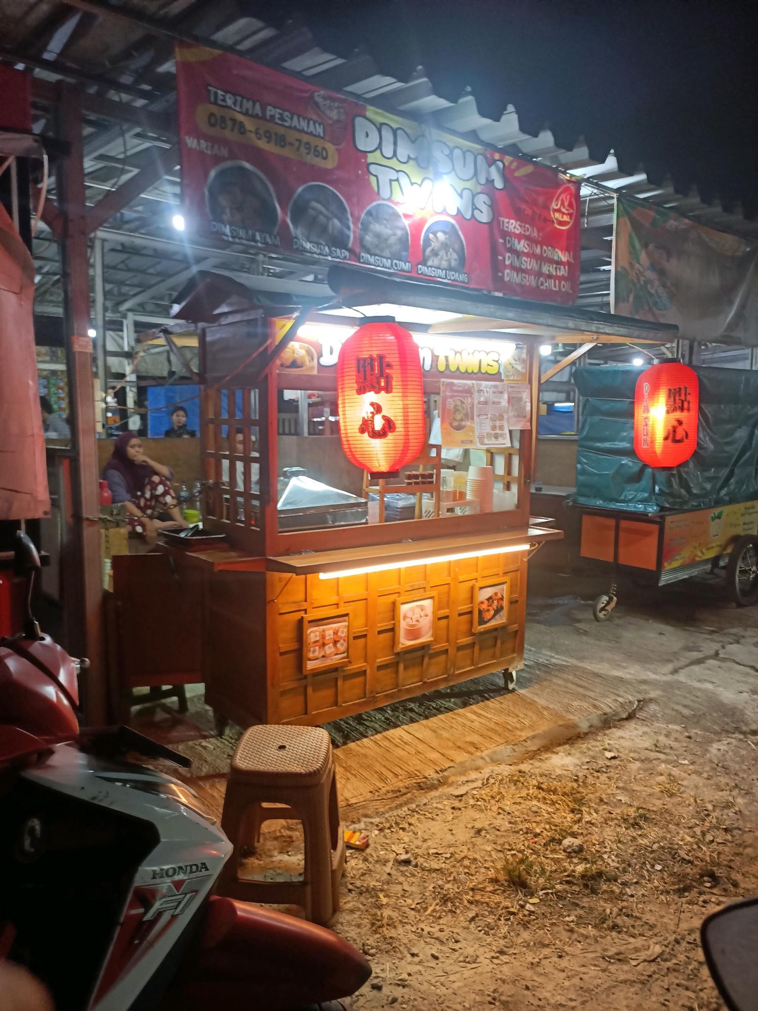 The image depicts an outdoor dim sum stand at night in Wanajaya, Indonesia. The stand is brightly lit, featuring a wooden cart with shelves displaying dim sum. Red paper lanterns with Japanese characters hang from the stand, adding to the ambiance. A red and white Honda motorcycle is parked in the foreground. In the background, a woman in a headscarf and polka dot pants is seated, observing. Another cart, covered with a green tarp, is parked to the right of the dim sum stand. The ground is covered with dirt and debris, suggesting a street-side location.