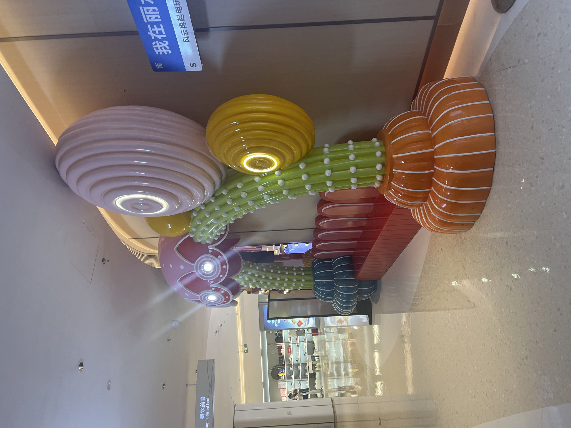 The image captures a retail interior in Liandu District, China, featuring a colorful, sculptural installation resembling stylized plants or fungi. The installation is composed of large, rounded elements in shades of pink, yellow, and orange, adorned with white dots and thin white stripes. A bright red bench is integrated into the base of the sculpture. The floor is a polished, light-colored terrazzo with reflective surfaces. In the background, storefronts are visible, displaying merchandise. A blue sign with Chinese characters and the letter "S" is mounted on a wall to the right of the sculpture. The overall environment suggests a shopping mall or a similar commercial setting, with artificial lighting and architectural elements like ceiling panels and light fixtures.