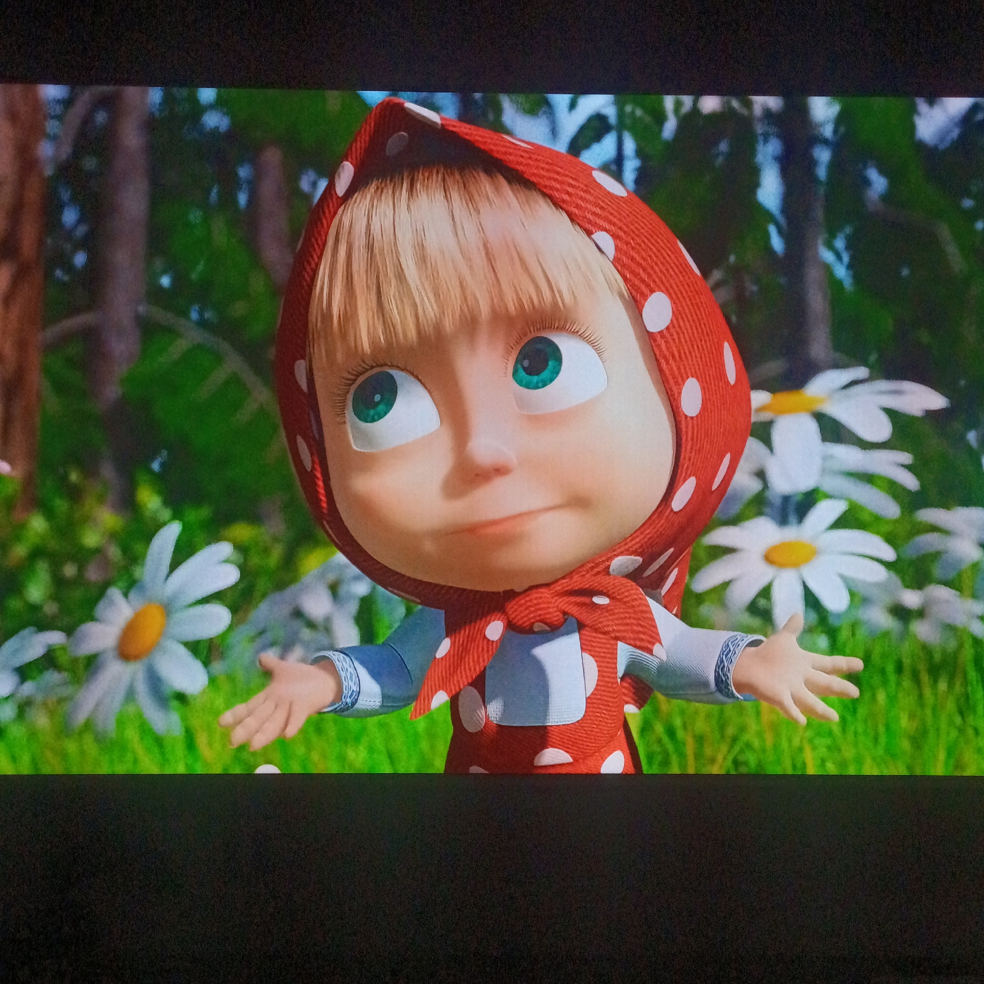 Here is a detailed description of the image:
The image shows a cartoon character, Masha, from the Russian animated television series "Masha and the Bear." She is a young girl with blonde hair, green eyes, and a mischievous expression. Masha is wearing a red headscarf with white polka dots and a red dress, also with white polka dots, over a white shirt with blue sleeves. Her arms are outstretched with open palms, as if in a gesture of surprise or welcome. The background features a lush green meadow with white daisies and a blurred forest backdrop.
I cannot verify that the character is in the location of Марковский, Russia, as it is a cartoon character and the background is ambiguous.
