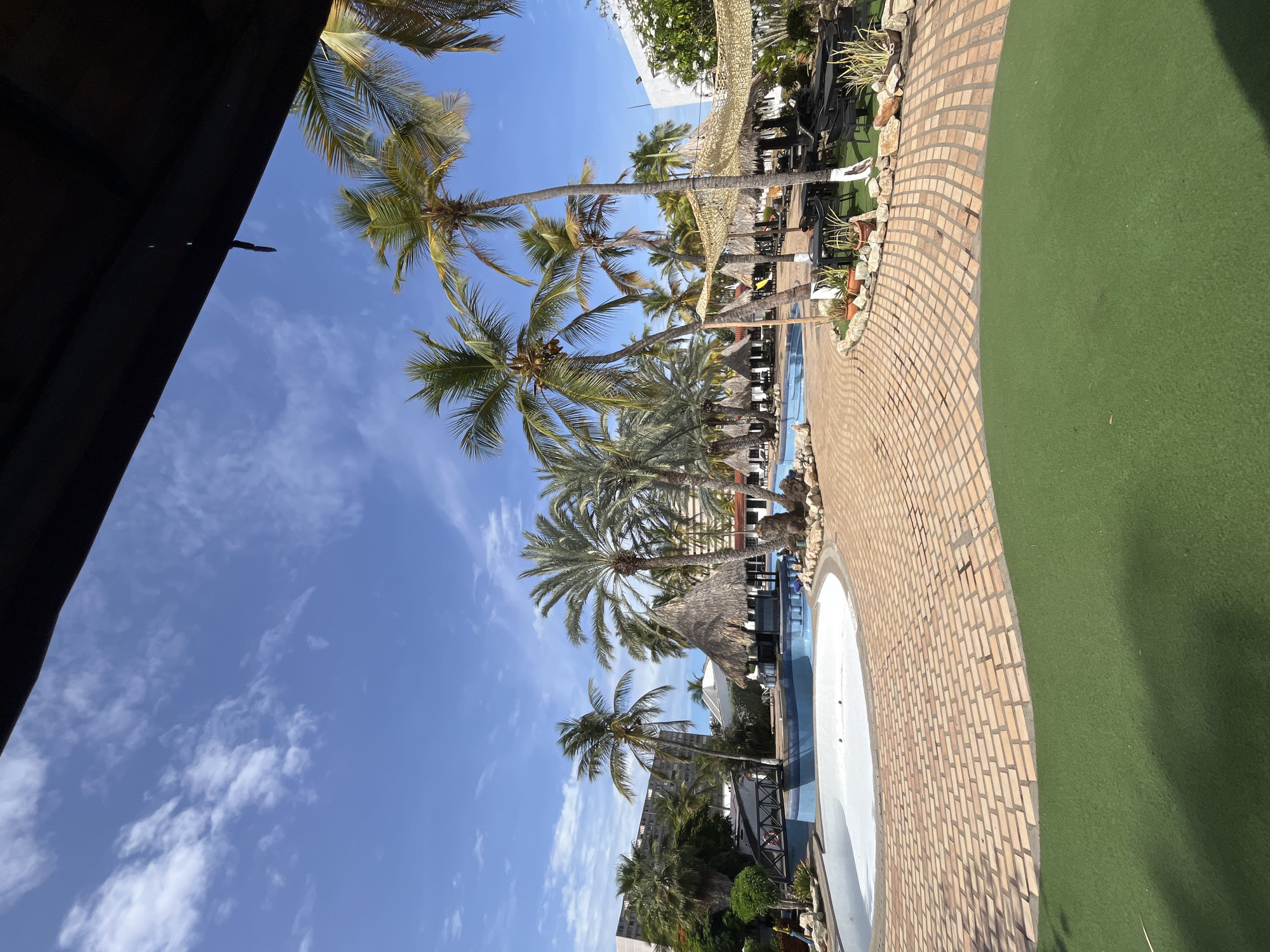 This image captures a serene outdoor scene at a tropical resort, likely located in Porlamar, Venezuela. The bright blue sky with scattered wispy white clouds fills the upper and left portions of the frame.
Numerous tall palm trees with lush green fronds are the dominant natural elements, extending from the mid-ground towards the sky, with some appearing to bear coconuts.
In the foreground and mid-ground, a swimming pool with a light blue edge and white interior is visible, appearing largely empty. It is bordered by a curved walkway paved with light brown, brick-patterned stones. Adjacent to this walkway is a broad expanse of vibrant green artificial turf or grass.
Around the pool and along the pathways, various resort amenities are set up. These include several thatched-roof structures (palapas or cabanas) providing shade, dark-colored lounge chairs, and tables, suggesting areas for relaxation. Potted plants and decorative rocks are interspersed along the edge of the paved path. A large building is partially visible in the background, surrounded by tropical foliage. A dark, possibly wooden, structure casts a shadow in the top-left corner, suggesting a roof overhang.
The scene is brightly illuminated by natural sunlight, casting clear shadows, indicating it is likely midday or early afternoon. The atmosphere appears peaceful and quiet, as no people are visible engaging in any activities. There is no visible text in the image.