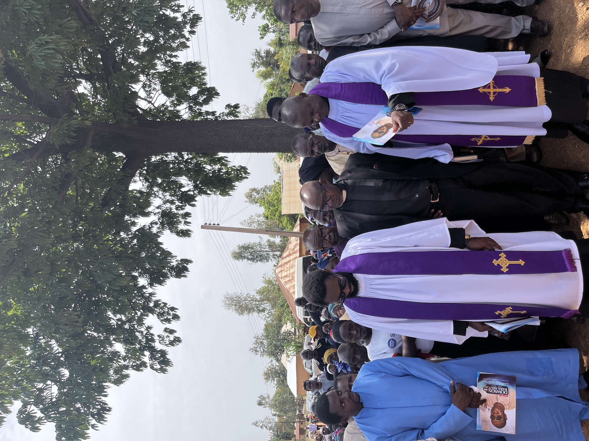 The image shows a procession of people, primarily clergy, in an outdoor setting that is possibly the city of Dogon Dinia, Nigeria. The group of clergymen are wearing traditional white robes with purple stoles adorned with gold crosses. Some individuals are holding what appear to be funeral programs featuring a portrait of a man named "Dowson Baba Akut". In the background, there is a residential area with trees, power lines and a corrugated iron roof visible. A large tree dominates the left side of the image. The ground appears to be dirt.