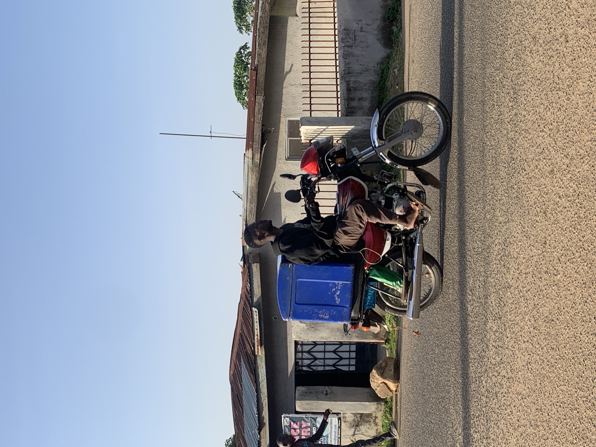 A man rides a red and silver motorcycle with a large blue cooler attached to the rear rack. He is wearing a black hoodie and brown pants. The motorcycle has a red helmet on its handlebars. The man is looking to his right. The motorcycle is on a paved road with a shadow cast on it. In the background, there is a grey building with a corrugated metal roof and a red metal fence. A person in a dark outfit is walking on the left side of the image. There is a large rock on the side of the road. The scene appears to be in Jos, Nigeria, given the context of similar images. The sky is clear and blue.