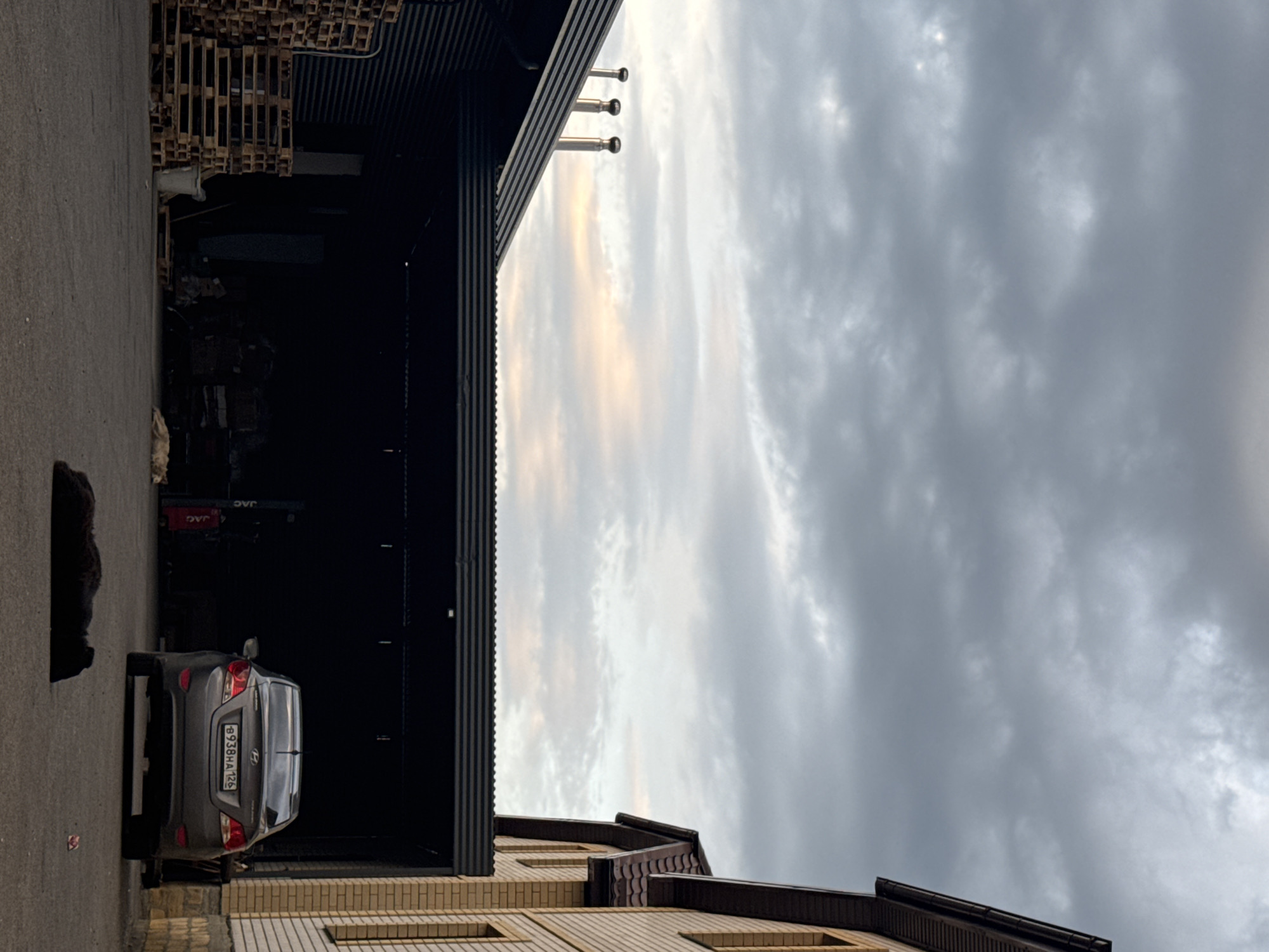 This is a vertical, low-angle shot of an outdoor industrial or storage area with a grey sedan parked in the foreground. The car is facing away from the camera, and its license plate reads "B938HA126". Behind the car, a dark, corrugated metal building casts a shadow. To the left of the building, stacks of wooden pallets are visible. Three exhaust pipes are mounted on the side of the building, extending upwards. The sky above is overcast with dramatic, grey clouds, hinting at twilight or a stormy day. The overall atmosphere is somewhat somber. The image appears to be taken in Mikhaylovsk, Russia, as indicated by the license plate format and the commonality of such industrial settings in that region. No people are visible in the image.