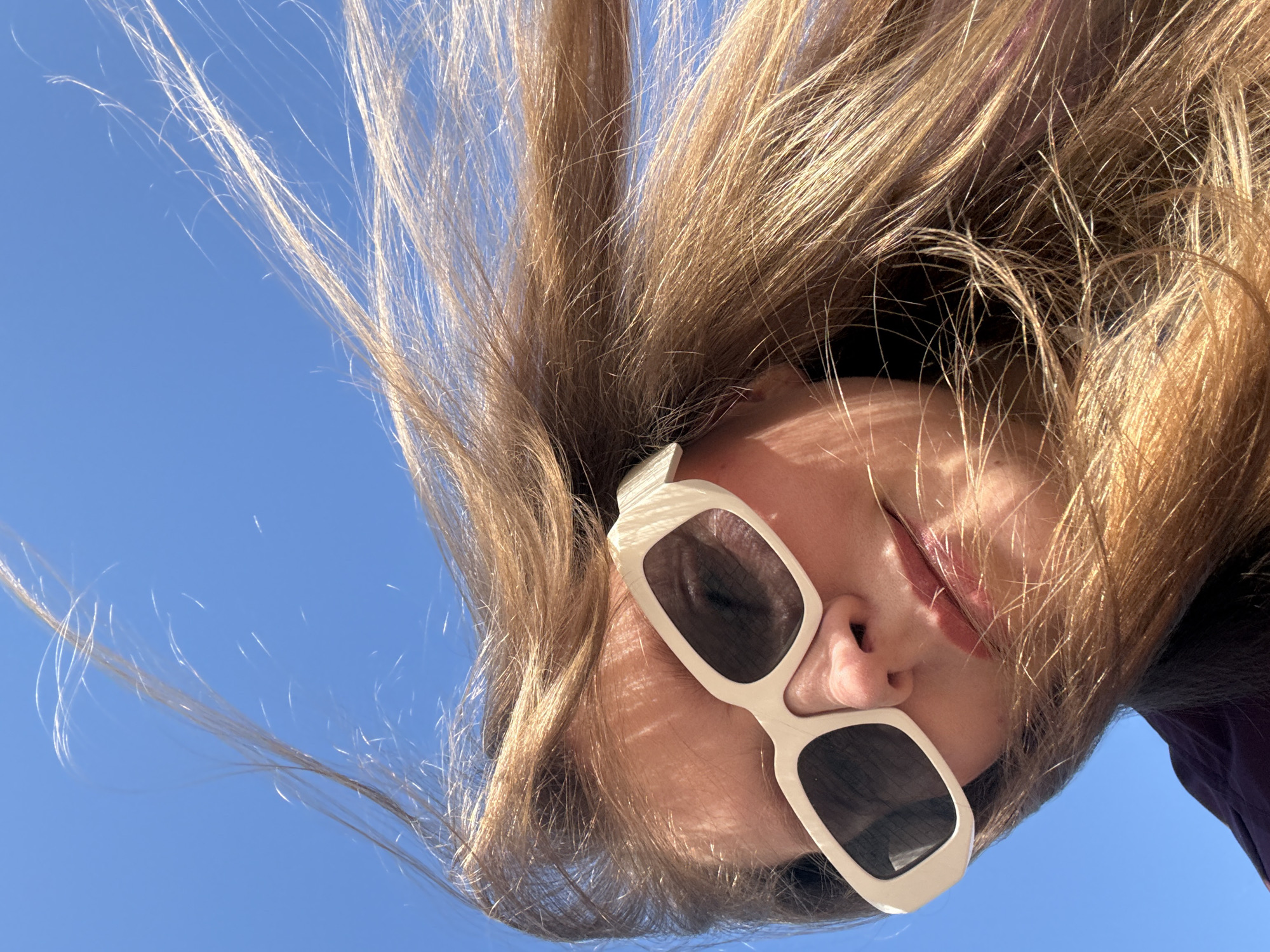 The image is an upward-facing shot of a person with light brown hair against a clear blue sky. The person is wearing white-framed sunglasses with dark lenses. The face is tilted towards the camera. The hair is tousled and spread out. The angle and position suggest a casual or playful pose. There is no information to determine that the image was taken in Izhevsk, Russia.
