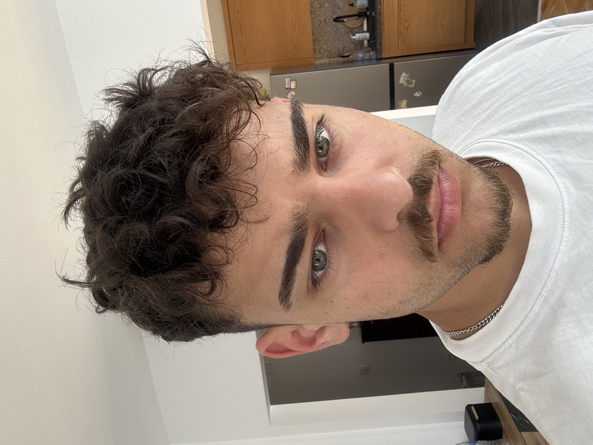 A close-up, portrait-style photograph shows a young man with dark, curly hair, light eyes, and a trimmed beard and mustache. He is wearing a white t-shirt and a thin silver chain necklace. The background is a domestic interior, including wooden cabinets, a stainless-steel refrigerator, and white walls. The location in the city of Troyan, Bulgaria cannot be determined from the image.