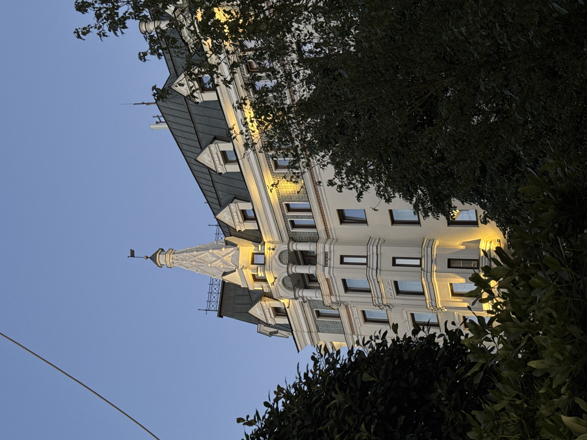 A multi-story building with a pale yellow facade and a dark green mansard roof is shown against a clear blue sky. The building features ornate architectural details, including a pointed spire with decorative trim and several dormer windows. Warm yellow lights illuminate sections of the building, suggesting it is evening or dusk. Lush green trees partially obscure the lower floors and right side of the building. A thin wire runs diagonally across the upper left portion of the image. The scene is consistent with the architecture found in Moscow, Russia, particularly in historical districts.