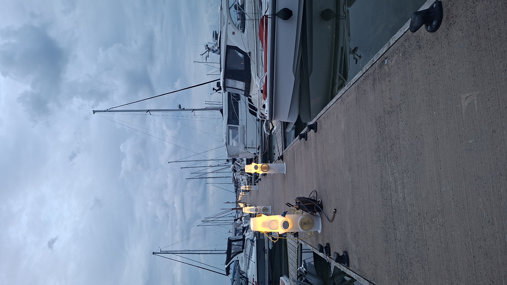 A concrete pier in a marina is lined with illuminated bollards, casting a warm glow on the overcast sky and surrounding sailboats. The pier is empty of people. The marina appears to be located in the city of Tallinn, Estonia, as evidenced by the characteristic architecture and signage that are typical of Estonian harbors. The water in the marina is calm and reflects the dim light.