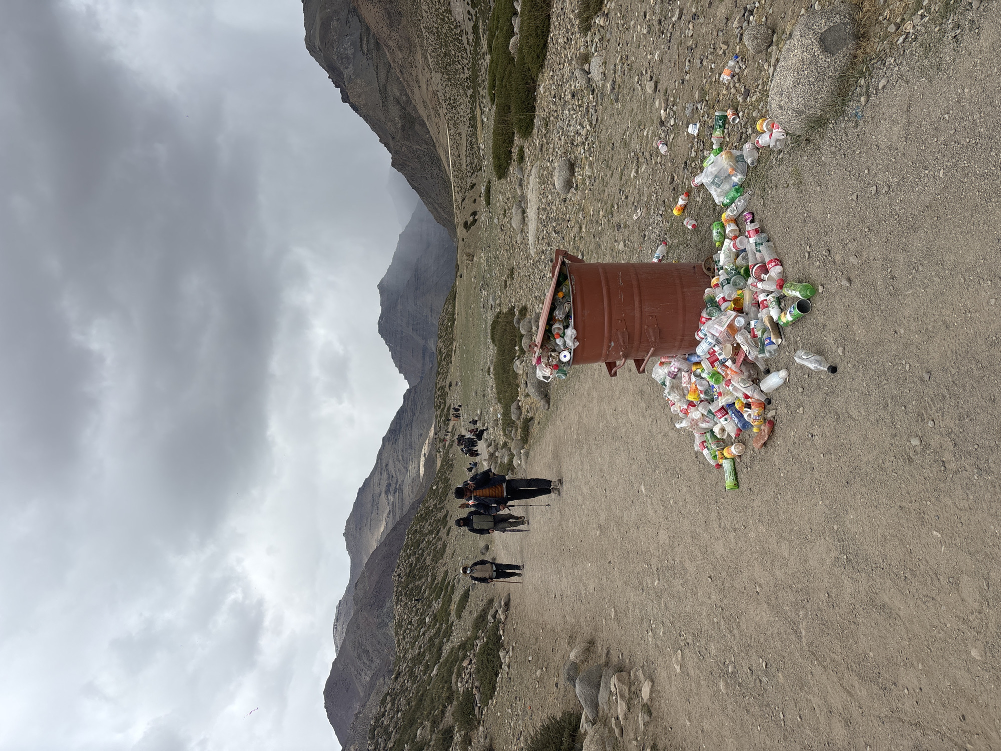 The image, rotated 90 degrees clockwise, presents a rugged, mountainous landscape under a dense, overcast gray sky. The scene unfolds along a wide, unpaved dirt path, and is contextually set in a mountainous region near Parga, China.
In the midground, a prominent, rusty brown metal barrel, serving as a waste bin, is severely overflowing. A substantial pile of discarded plastic bottles and aluminum cans has spilled out onto the ground beside it, highlighting a significant issue with litter or waste management in this natural setting. Visible brands on the refuse include "Coca-Cola," "Sprite," and "Fanta."
Several individuals are seen walking along the dirt path, which is composed of packed earth and small rocks. They appear to be hikers or trekkers, with some carrying backpacks and utilizing walking poles, indicating an active journey through the terrain.
The background is dominated by majestic, barren mountains that rise steeply, their rugged forms fading into the cloudy distance. Sparse green vegetation dots the lower slopes, providing a subtle contrast to the predominantly rocky and earthy tones of the environment. The uniformly gray sky casts a soft, diffused light over the entire scene, creating a somewhat somber atmosphere. The image captures the striking juxtaposition of natural beauty and human environmental impact.