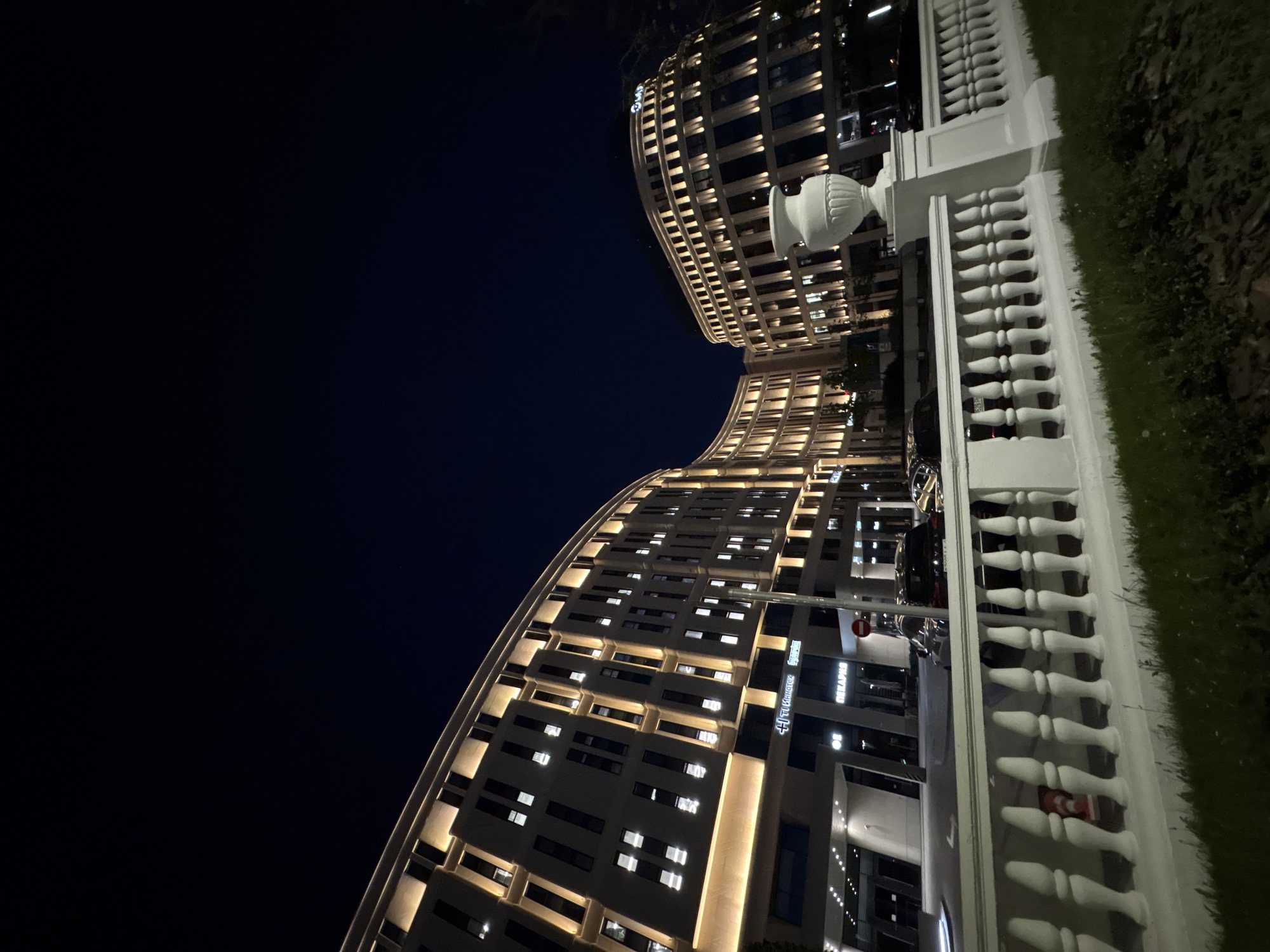 At night, a low-angle shot captures a modern architectural scene in Minsk, Belarus. The upper left of the image is dominated by a dark sky. Two curved, multi-story buildings, illuminated with numerous lighted windows, rise from the bottom of the frame. A white balustrade runs from right to left along the foreground, with a patch of green grass bordering it on the right. Cars and signs are visible at the base of the buildings.