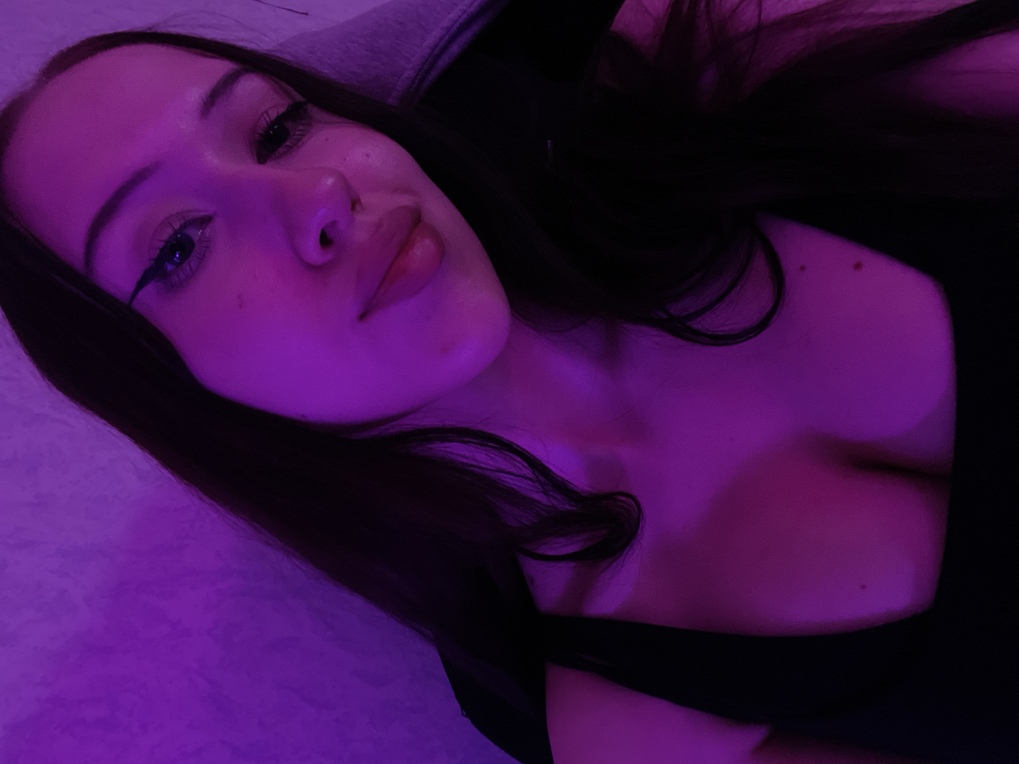 The image is a selfie of a young woman with long, dark hair, bathed in purple lighting. She is lying down, with her head tilted back and her body angled towards the viewer. Her eyes are open and she is looking directly at the camera with a neutral expression. She is wearing a black top that reveals her décolletage. Her makeup includes winged eyeliner and lip gloss. The background is a soft, textured surface, possibly a bed or sofa, illuminated by the purple light. The overall mood is intimate and relaxed. The image is taken in Votkinsk, Russia. There is no visible text in the image.