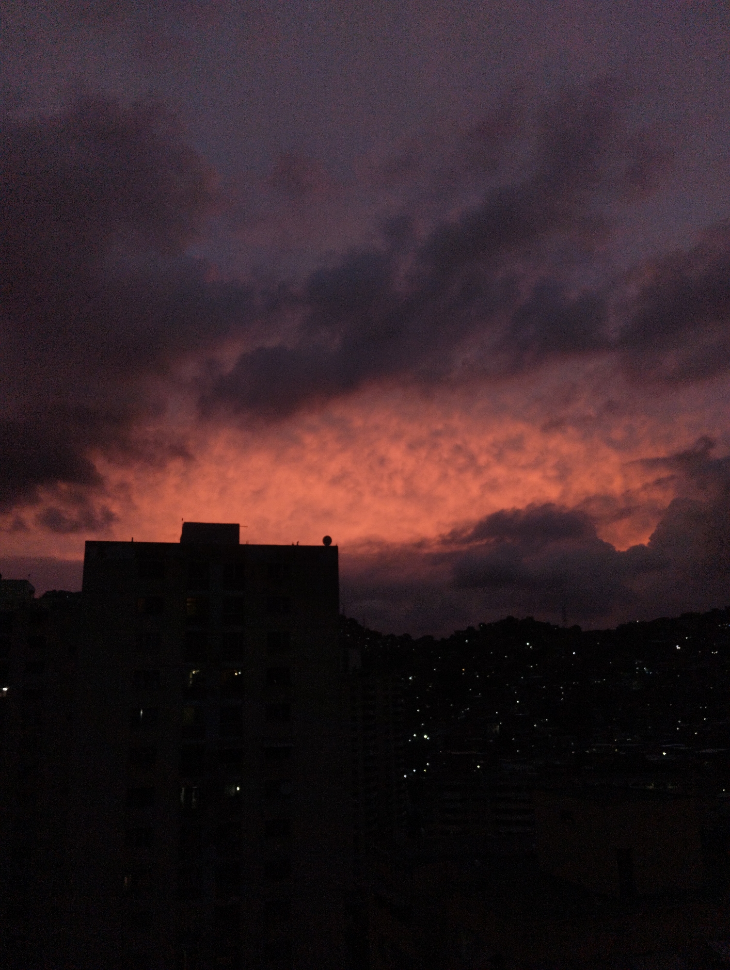 The image captures a night view in Caracas, Venezuela, featuring a skyline dominated by a tall, dark building silhouetted against a dusky sky. The sky exhibits a gradient of colors, transitioning from a vibrant orange near the horizon to dark purple clouds above. On the right side of the frame, a hillside dotted with numerous small lights suggests a residential area at night. Other buildings and structures, also in silhouette, are visible in the foreground. There are no people visible in the image.