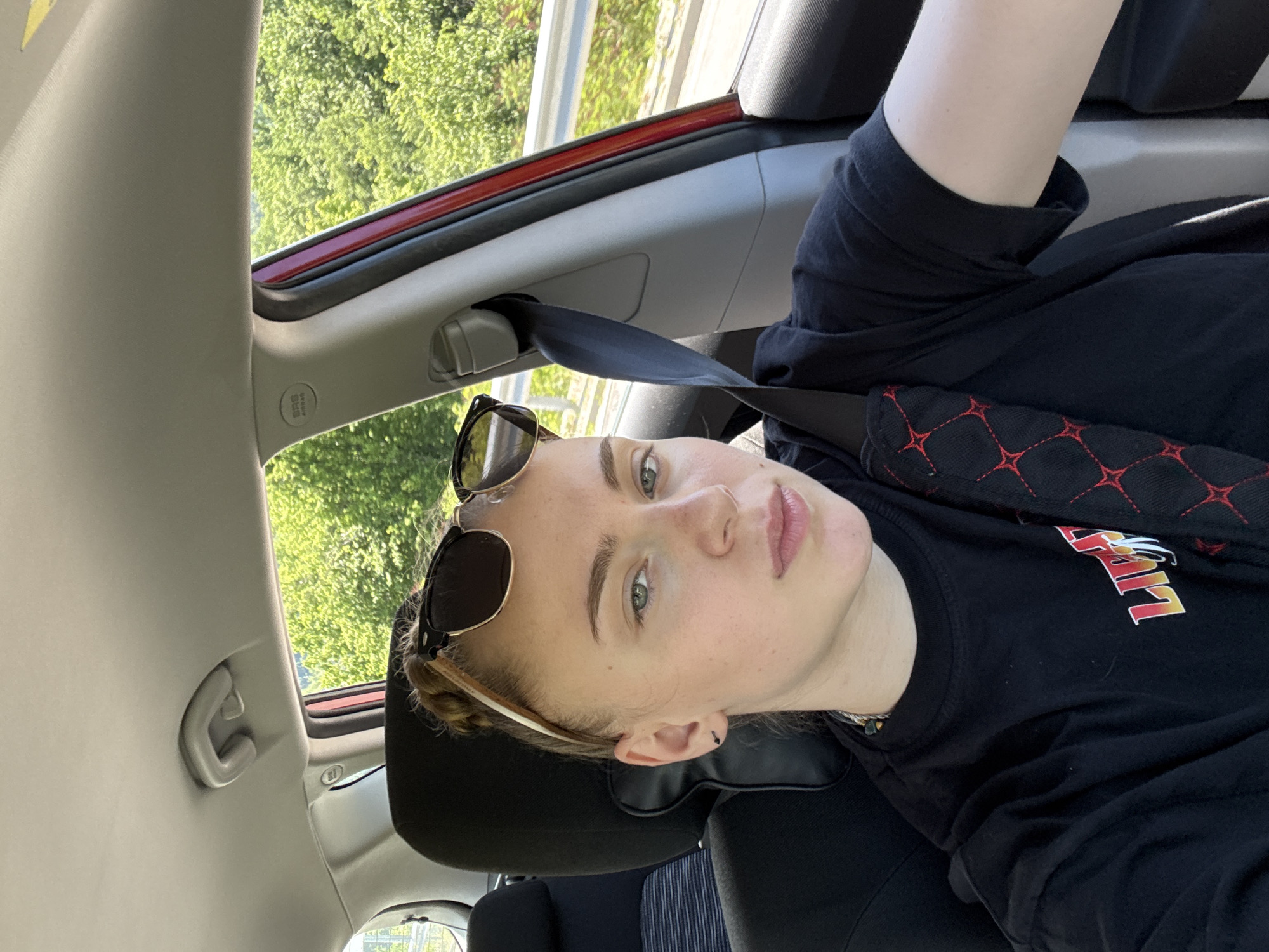 A medium shot captures a young person with blonde hair pulled back, wearing sunglasses perched on their head, and a black t-shirt with a graphic design on it. They are seated in the passenger seat of a car, indicated by the visible seatbelt and car interior. The person is looking towards the camera, with a neutral expression. Outside the car, lush green trees and vegetation are visible, suggesting a rural or suburban setting. The lighting suggests it is daytime. The image does not provide any specific clues that would place the scene in Gabrovo, Bulgaria.