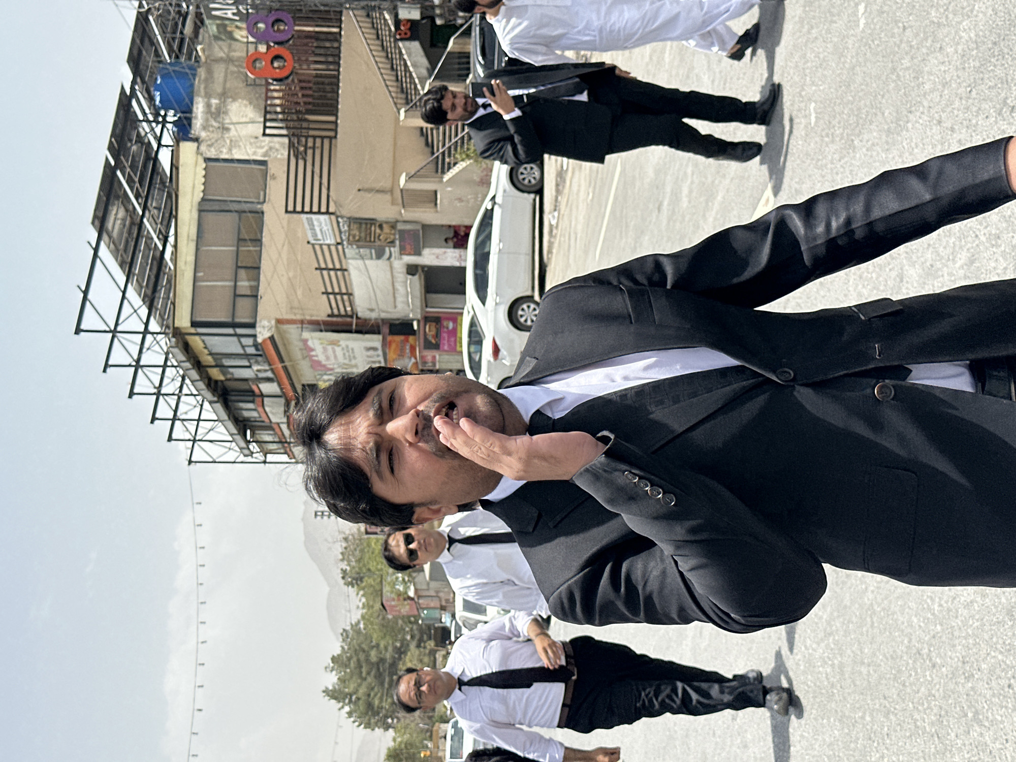 In Quetta City Tehsil, Pakistan, a man in a black suit and tie is captured mid-shout, his hand cupped near his mouth, on a street during the daytime. He is the primary subject, positioned in the foreground with a shallow depth of field focusing on him. Behind him, several other men in similar formal attire are walking. One man in a white shirt and black tie is walking slightly behind and to his left, while another in a white shirt and black tie is further back and to the left. Another man in a black suit is visible to the right, looking at his phone. Further in the background, a man in a white shalwar kameez is walking away from the camera. The street appears to be paved, with white cars parked on the right side. Buildings line the street, some with awnings and signage, including a prominent sign with the numbers "86" in red and purple. The sky is overcast, and trees and mountains are visible in the distance.