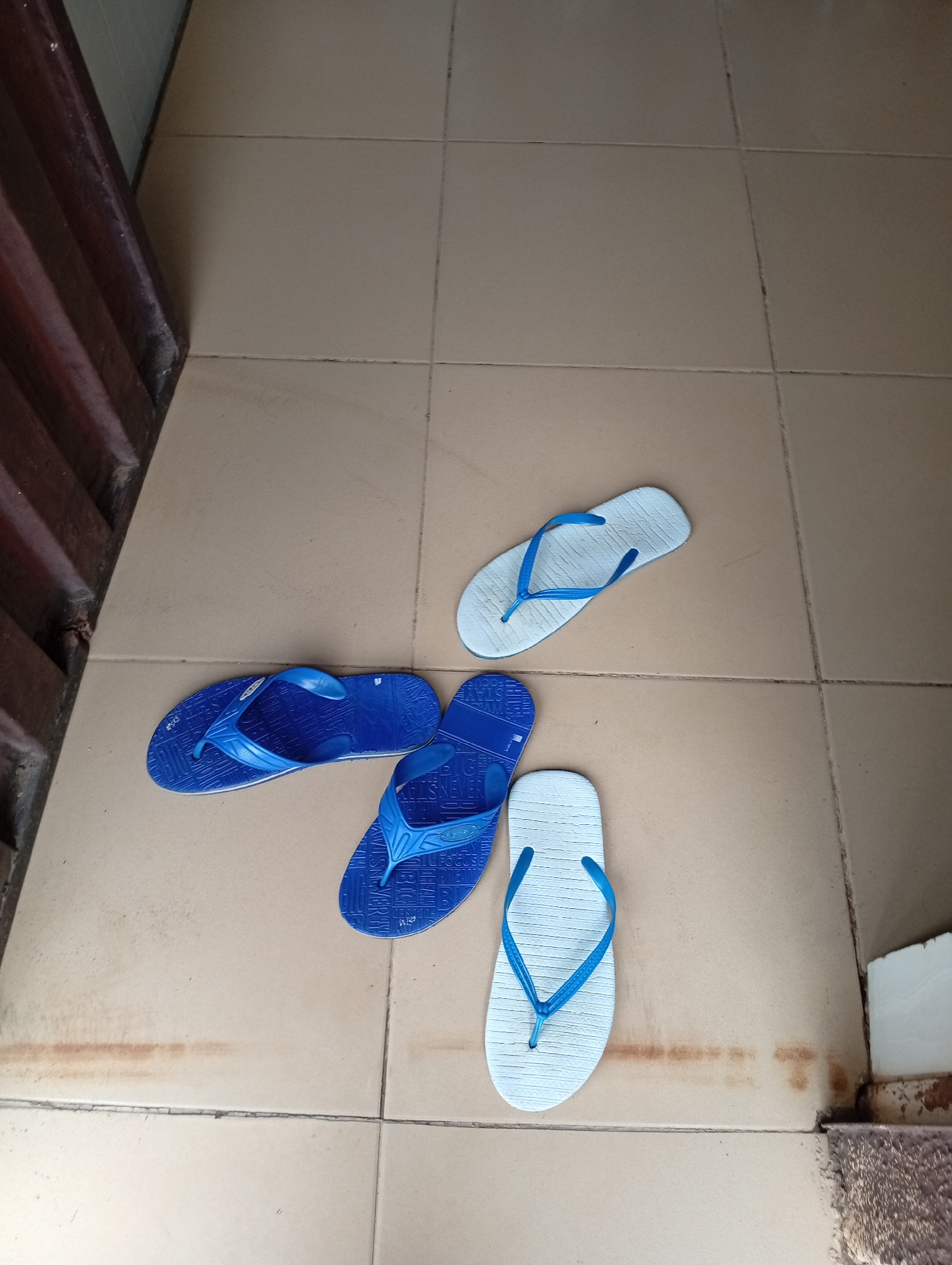 Four flip-flops are displayed on a tiled floor. Two flip-flops are blue, and two are light blue/white. One blue flip-flop is on the left side of the image, with the sole facing upwards and the straps forming a V-shape. To its right and slightly below it is another blue flip-flop, also with the sole facing upwards and the straps forming a V. Above and to the right of these two blue flip-flops is a light blue/white flip-flop, sole upwards, straps forming a V. Below this light blue/white flip-flop and slightly to the right is another light blue/white flip-flop, sole upwards, straps forming a V. The floor is made of square, light brown tiles with dark grout lines. To the left of the tiles is a dark brown, corrugated surface, possibly a door or wall. The scene appears to be outdoors or in an open-air entryway. This scene is located on the floor of a building in Rafin Par, Nigeria.