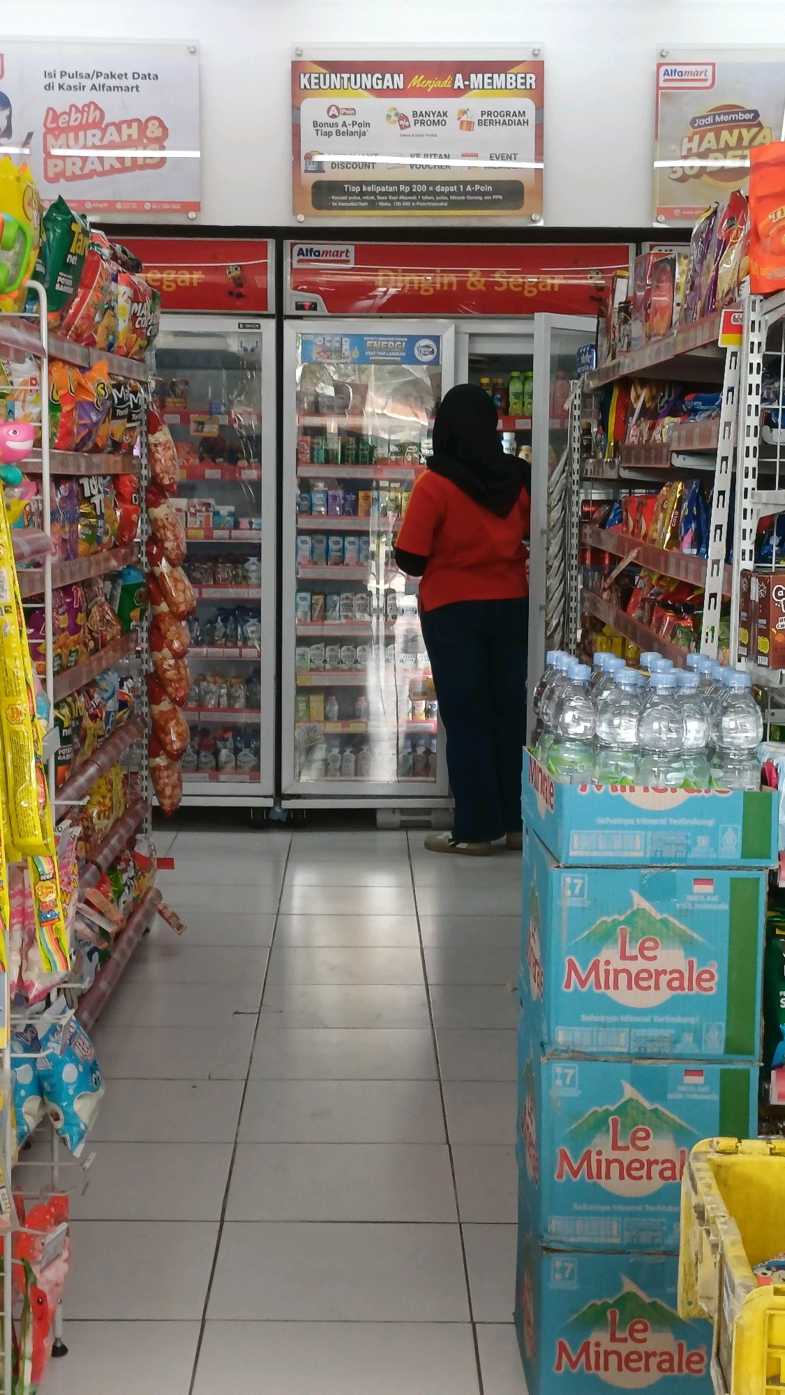The image shows the interior of an Alfamart convenience store in Tengahan, Indonesia. The perspective is from the aisle, looking towards the back of the store. On the left, shelves are stocked with various packaged snacks and goods. In the center, there are two large refrigerated display cases filled with beverages. A person wearing a red shirt and dark pants, with a black hijab, is standing with their back to the camera, looking into the refrigerated case. To the right, more shelves are filled with products, and a stack of blue boxes labeled "Le Minerale" water is visible in the foreground. The floor is tiled with light-colored square tiles. Above the refrigerated units, promotional posters are displayed on the wall. One poster announces "Keuntungan Menjadi A-Member," detailing member benefits. Another poster says "Jadi Member HANYA SOPE." The overall impression is a clean and well-stocked convenience store.