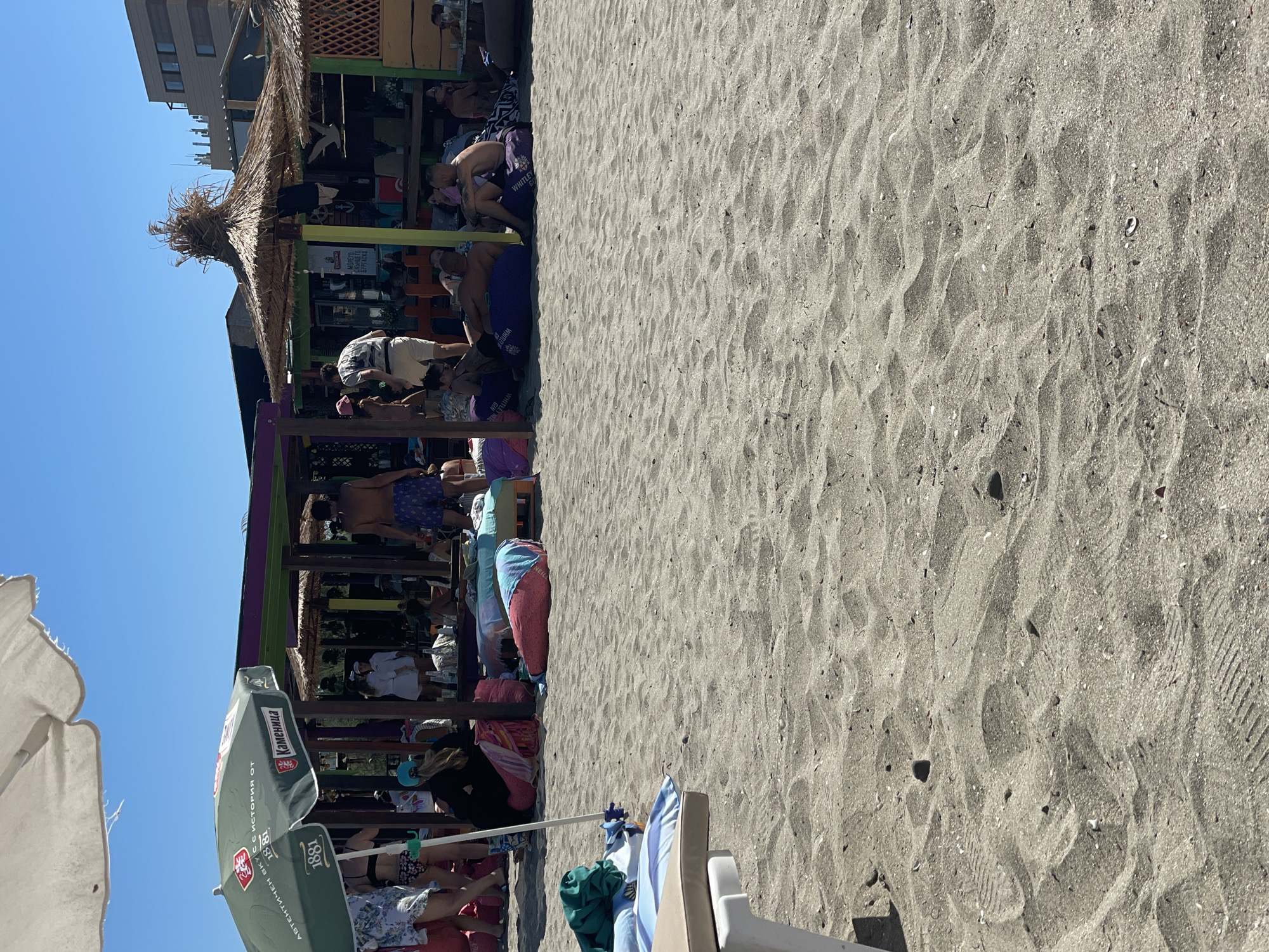 A group of people are relaxing on a sandy beach under umbrellas and a thatched-roof structure, likely a beach bar. The scene is located in Pomorie, Bulgaria, a popular seaside resort town. The foreground is dominated by sand with some scattered shells and debris, along with a portion of a lounge chair. In the middle ground, several individuals are seated on large beanbag chairs or lying on the sand. Some are partially obscured by beach umbrellas and a colorful, open-sided structure that appears to be a bar or seating area. The background shows more beachgoers, vegetation, and buildings, suggesting a developed area. The sky is clear and blue.