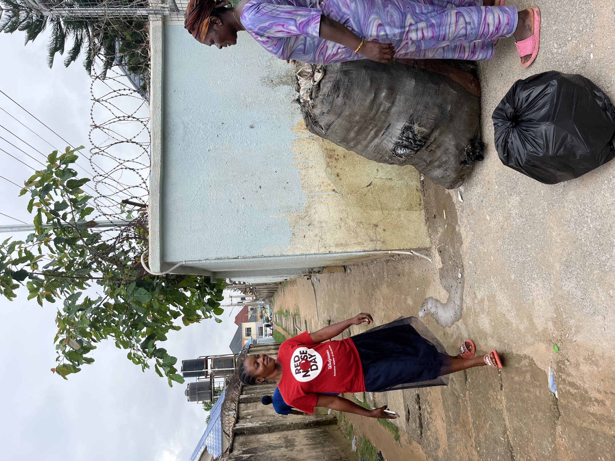 In Abuja, Nigeria, two women stand in an alleyway. The woman on the left, wearing a red t-shirt that reads "RED NOSE DAY" and a navy blue skirt, walks towards the camera, holding a phone in her left hand. The woman on the right, dressed in a purple and white patterned dress and a headscarf, faces the woman in red and looks away from the camera. She wears a gold bracelet on her right wrist and pink sandals. In front of her, a large, dark, burlap sack is filled with trash, and a black plastic trash bag lies on the ground beside it. The alleyway has a concrete path with some debris. To the left, a fence with barbed wire lines the alley, with trees and buildings visible in the background. The sky is overcast.