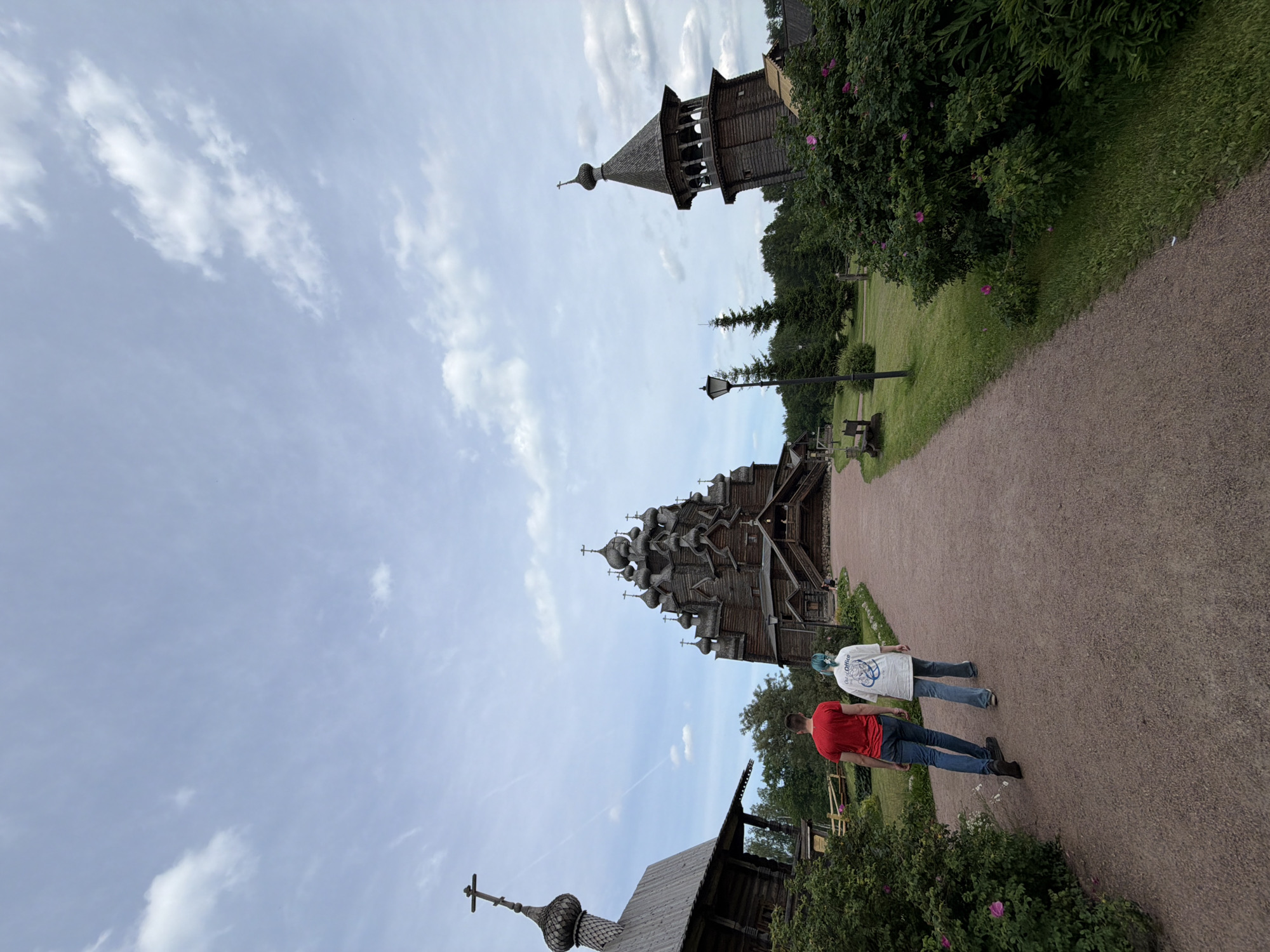 The image captures the Church of the Transfiguration, a wooden church located in the city of Rabochii, Russia, on Kizhi Island in the Republic of Karelia. In the foreground, a man wearing a red t-shirt and blue jeans walks on a gravel path, followed by a person in a white t-shirt and blue jeans with light blue hair. They are walking away from the camera, towards the imposing wooden church. The church is a multi-domed, intricately carved wooden structure, characteristic of traditional Russian architecture. To the left of the church, another smaller wooden building with a dark roof and a cross on top is visible. To the right, a wooden bell tower, also topped with a cross, stands at an angle. The surroundings are a lush green landscape with trees and bushes, some of which are in bloom with pink flowers. The sky is partly cloudy, with a pale blue hue. The overall scene depicts a peaceful, architectural and natural setting.