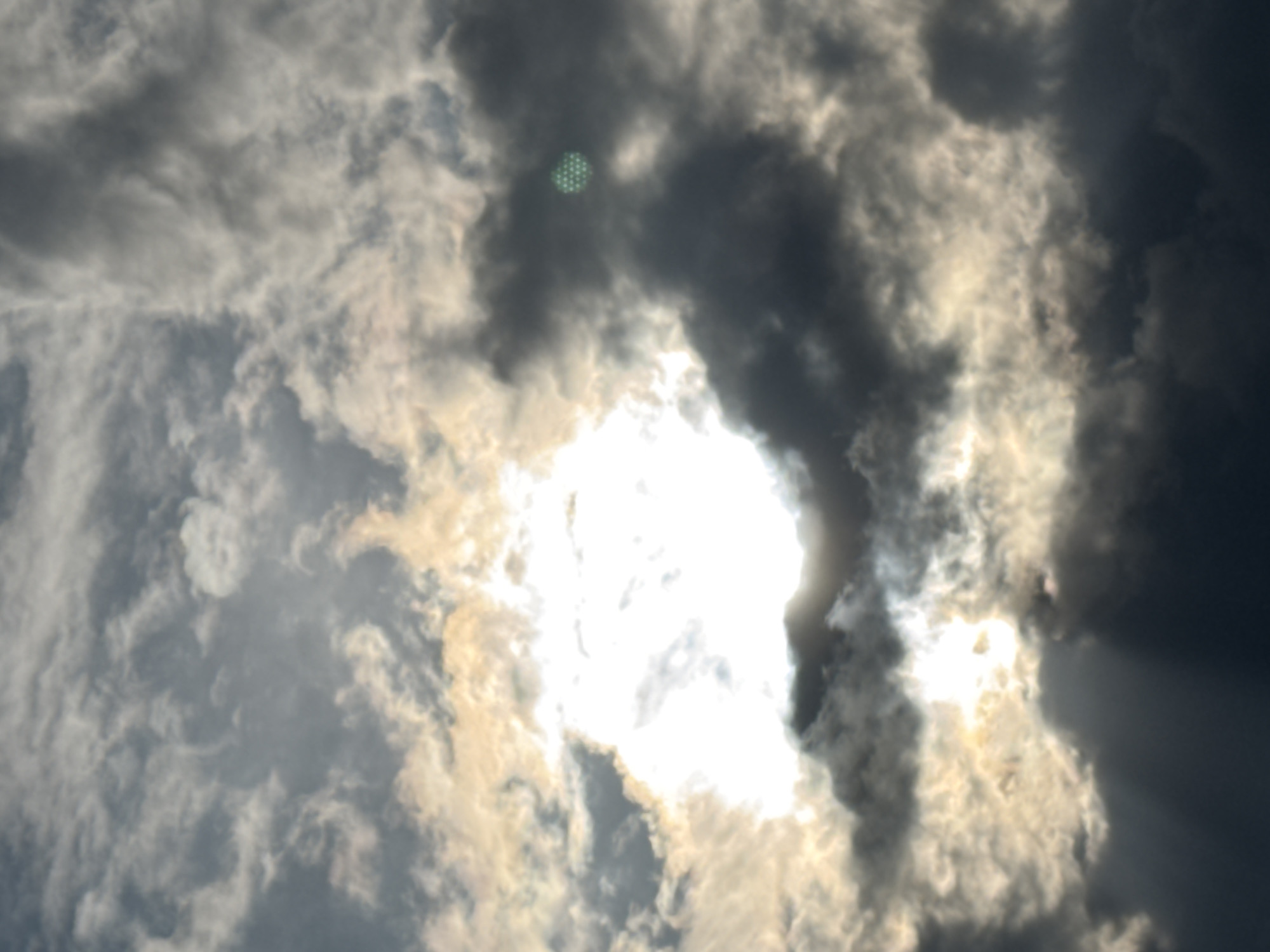 A dramatic sky with dark, ominous clouds and bright sunlight breaking through. A circular green object with a honeycomb pattern is visible in the upper right quadrant. The sun, partially obscured by clouds, casts rays of light downwards, creating a spotlight effect. The scene is likely in Sary-Oy, Kyrgyzstan, given the context.