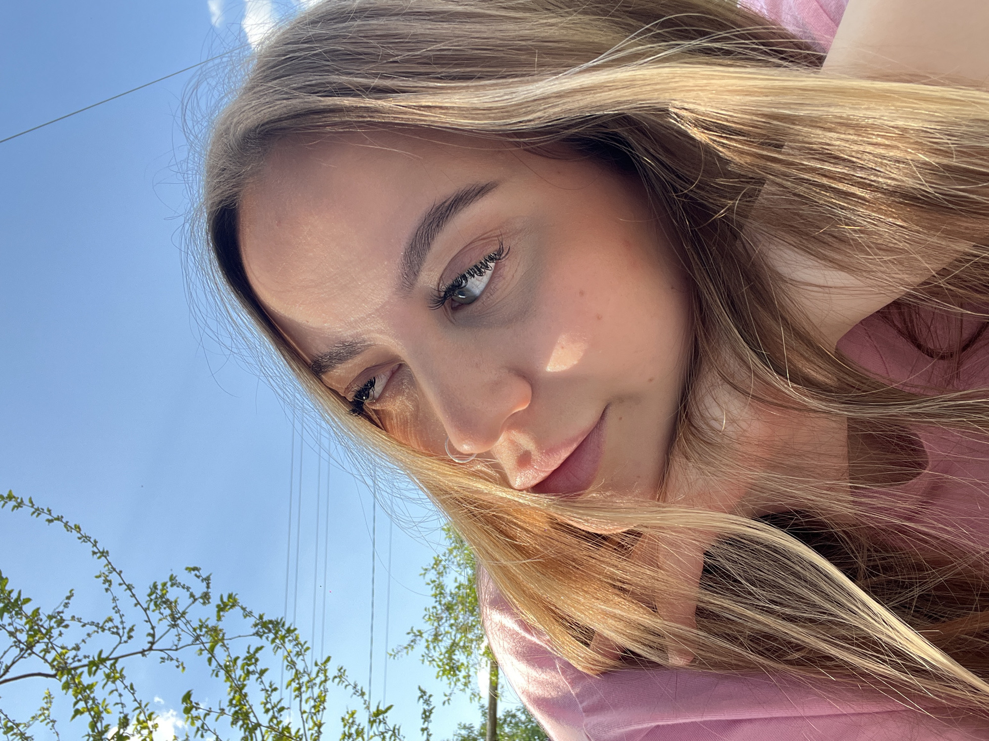 A young woman is centered in the frame with a slightly tilted head and face towards the camera. The backdrop reveals a clear blue sky dotted with sparse clouds, foliage from nearby trees, and parallel power lines. The subject's hair is long and blond, cascading around her face. Her makeup is natural, with a hint of mascara. She wears a pink top and has a nose piercing.