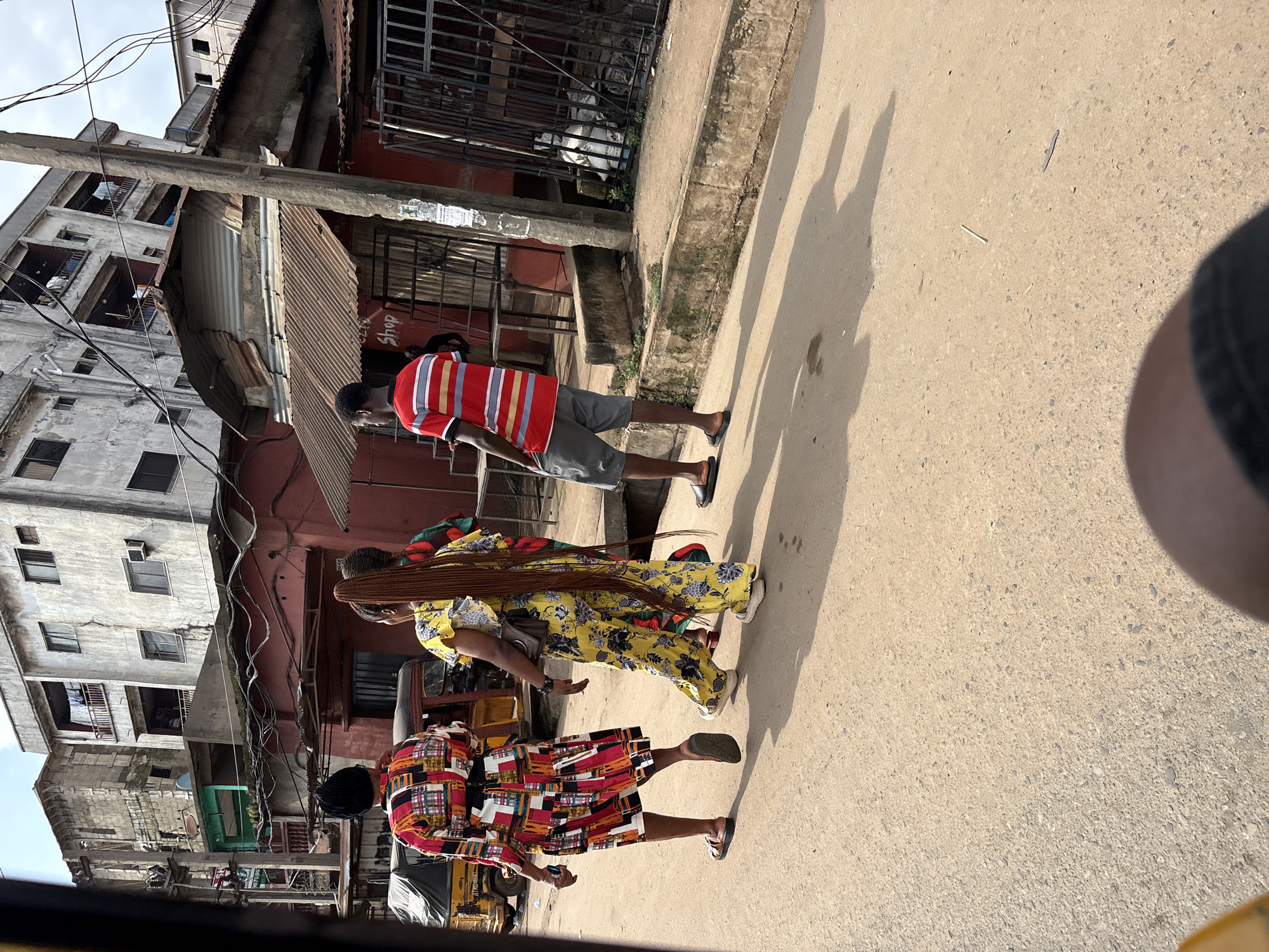 The image depicts a street scene in Onitsha, Nigeria, characterized by a mix of residential and commercial buildings. In the foreground, three individuals are walking on a paved street. The person on the left, viewed from the back, wears a colorful, patterned dress. Next to them, a person with long braided hair wears a bright yellow jumpsuit with a floral pattern. To the right, a man in a red and white striped shirt and grey shorts walks ahead. Behind them, a yellow auto-rickshaw is partially visible on the left side of the frame. The background features multi-story buildings with weathered facades, some appearing to be under construction or in disrepair, and a corrugated metal roof structure over a storefront labeled "Shop." Electrical wires crisscross the sky above. The ground is a dusty, paved surface with visible shadows cast by the people and buildings, suggesting a sunny day. The overall impression is a bustling urban environment in Nigeria.