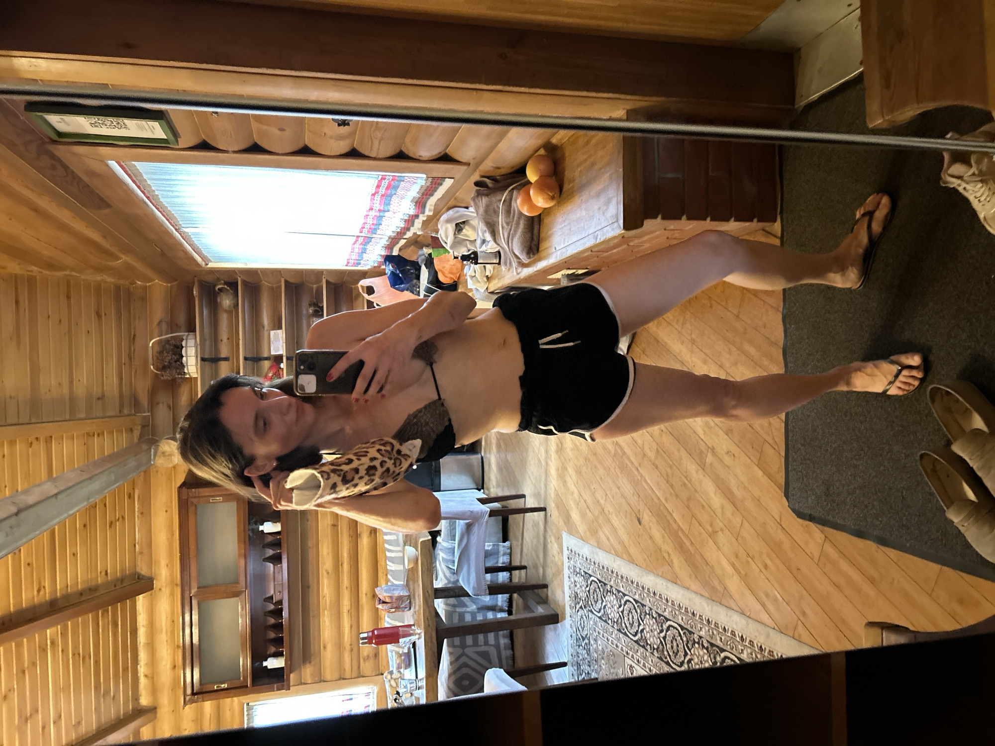 A woman is taking a selfie in a mirror. She is wearing a black bikini top and black shorts with white trim. She is holding a leopard-print pillow in her left hand and a cell phone in her right. The reflection shows a log cabin interior, including a window, dining table, rug, and fireplace. There is no indication of Khimki, Russia.