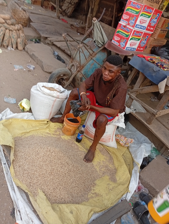 A man sits barefoot among bags of grain on a yellow tarp in an outdoor market setting. He wears a striped brown shirt and red shorts, adorned with gold jewelry. A bucket and soda bottle sit nearby. Boxes of Indomie noodles are stacked on a table behind him. The area around him appears cluttered with various items, including a cart, sacks, and scattered debris, suggesting a busy commercial environment.