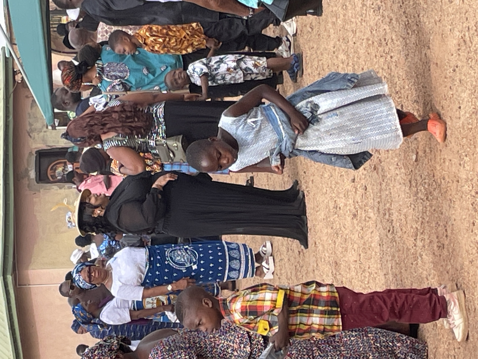 A group of people are outdoors, seemingly gathered for an event. The location is likely in Jos, Nigeria (based on cultural cues in clothing). The ground is dirt. The people are dressed in a variety of clothing, including traditional African attire. There are several adults and children visible. Some individuals are wearing hats. One child is wearing a light blue dress and orange shoes. Another child is wearing a plaid shirt and maroon pants.