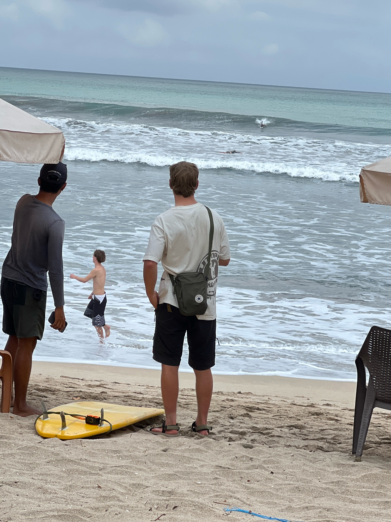 Two men stand on a sandy beach in Kuta, Indonesia, facing the ocean. One man, on the left, wears a grey long-sleeved shirt and dark shorts, with a dark baseball cap. The other man, on the right, wears a white t-shirt with a graphic print and dark shorts, and a green crossbody bag. In the shallow water between them, a shirtless young man in black and white patterned shorts runs into the waves. Further out in the ocean, a surfer is riding a wave. A yellow surfboard lies on the sand in the foreground. Beach umbrellas are visible on the left and right sides of the frame, and a dark plastic chair is situated to the right. The sky is overcast, and the ocean is a muted turquoise with white-capped waves.