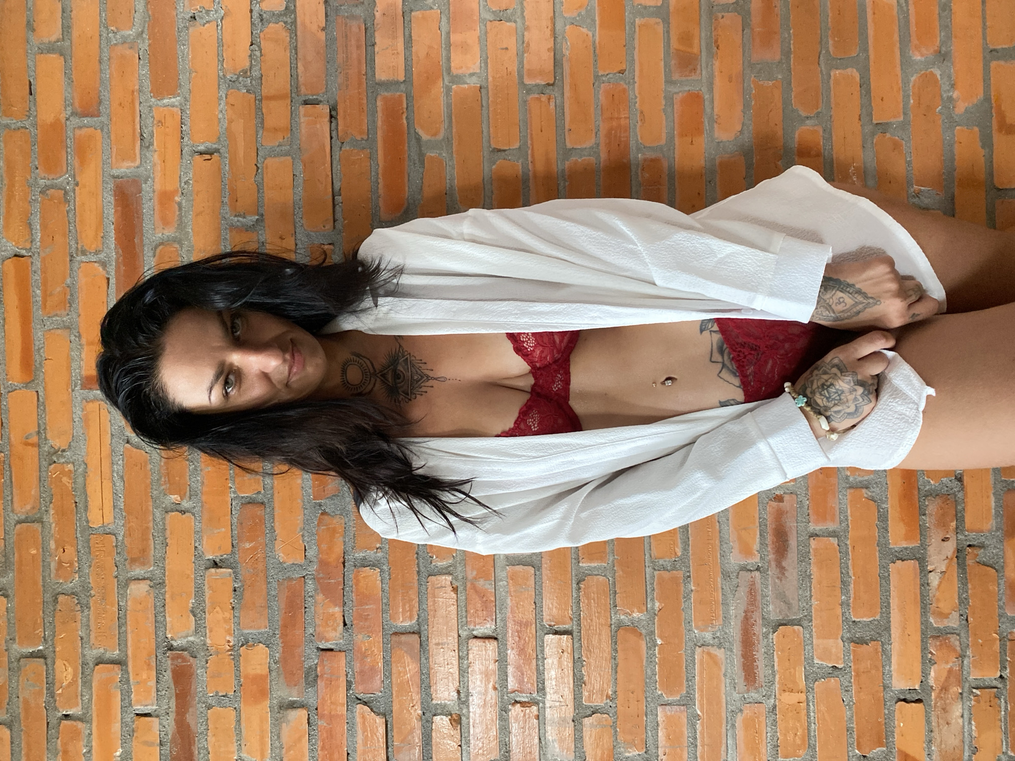 A woman with long, dark hair is depicted lying horizontally against a rustic red brick wall. She looks directly at the viewer with a slight smile and confident expression. She is wearing a red lace bra and a white, unbuttoned shirt or robe that is loosely draped over her body.
Notable details include several tattoos: a mandala-like design on her chest, intricate patterns on her hands and arms, and possibly more on her hip/thigh area, partially visible beneath the white shirt. She also has a belly button piercing and wears bracelets on her right wrist. The background consists of a textured wall made of reddish-orange bricks with grey mortar. The lighting is soft, creating a focused portrait. The setting is likely Mũi Né, Vietnam, as indicated by the provided context. There is no visible text in the image.