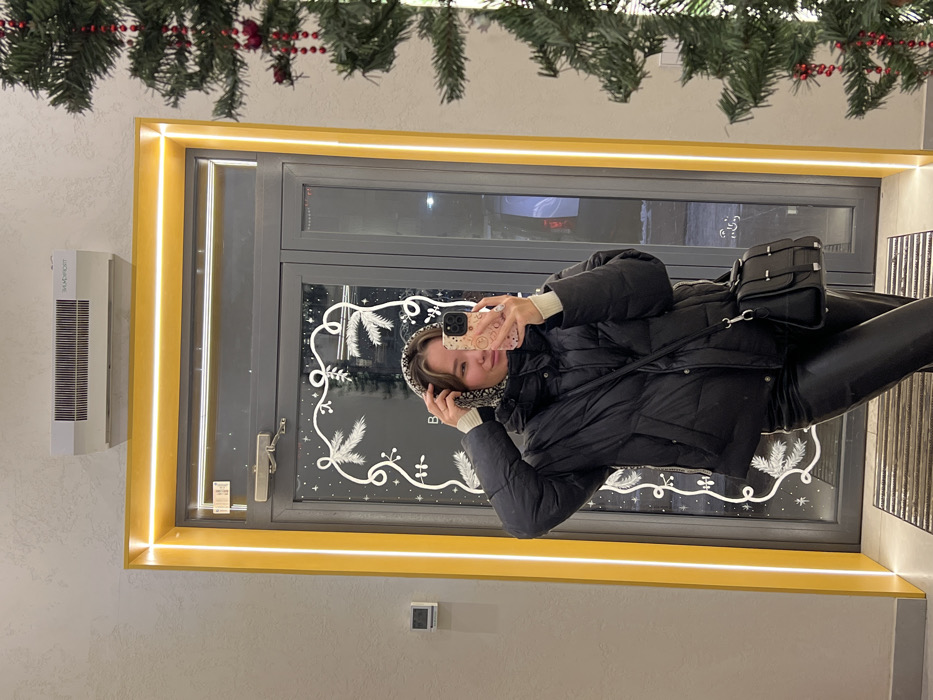 A young adult female is depicted taking a selfie within a building entrance in Krasnoyarsk, Russia. She stands approximately chest-high in the frame, angled slightly to her left, holding a light-colored smartphone with a decorated case in her right hand towards the reflective surface to capture the image. Her left hand is positioned near a patterned head covering.
She wears a black quilted puffer jacket, black form-fitting trousers with a glossy finish, and a light-colored long-sleeved top visible at her neck and cuffs. A black shoulder bag with a silver-colored clasp mechanism hangs across her body. She has dark hair, a light complexion, and a faint smile.
The background consists primarily of a double-paned glass door, framed by a prominent yellow border with integrated LED strip lighting, emanating a warm glow. The glass is adorned with white, winter-themed decals depicting stylized foliage and snowflakes. Through the reflection in the door, interior elements are visible, including indistinct light sources and structural elements. A dark-colored, ribbed doormat is visible at the bottom of the frame.
Above the door, mounted on a light grey textured wall, is a white wall-mounted air curtain unit bearing the Cyrillic inscription "ЭНЕРГОФОРТ" (ENERGOFORT), a Russian brand. To the far right, an artificial Christmas tree, decorated with red beaded garlands, partially enters the frame. To the left of the door, on the grey wall, a small dark square object, possibly a thermostat or sensor, is mounted.
