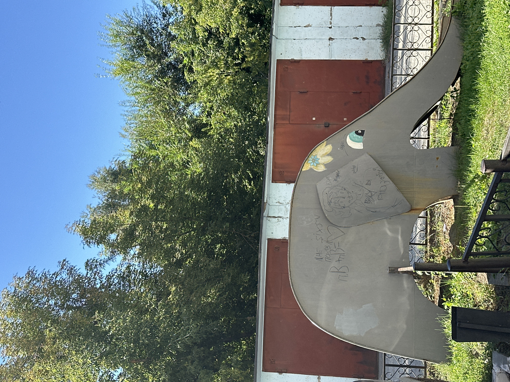 A metal elephant sculpture, part of a children's playground, is positioned in front of a row of garage doors in Biysk, Russia. The sculpture is grey and shows signs of graffiti. A yellow flower is painted on its head, and its eye is a turquoise circle. The background consists of green trees and a clear blue sky. The elephant sculpture is likely located in a residential area, as indicated by the presence of garages.