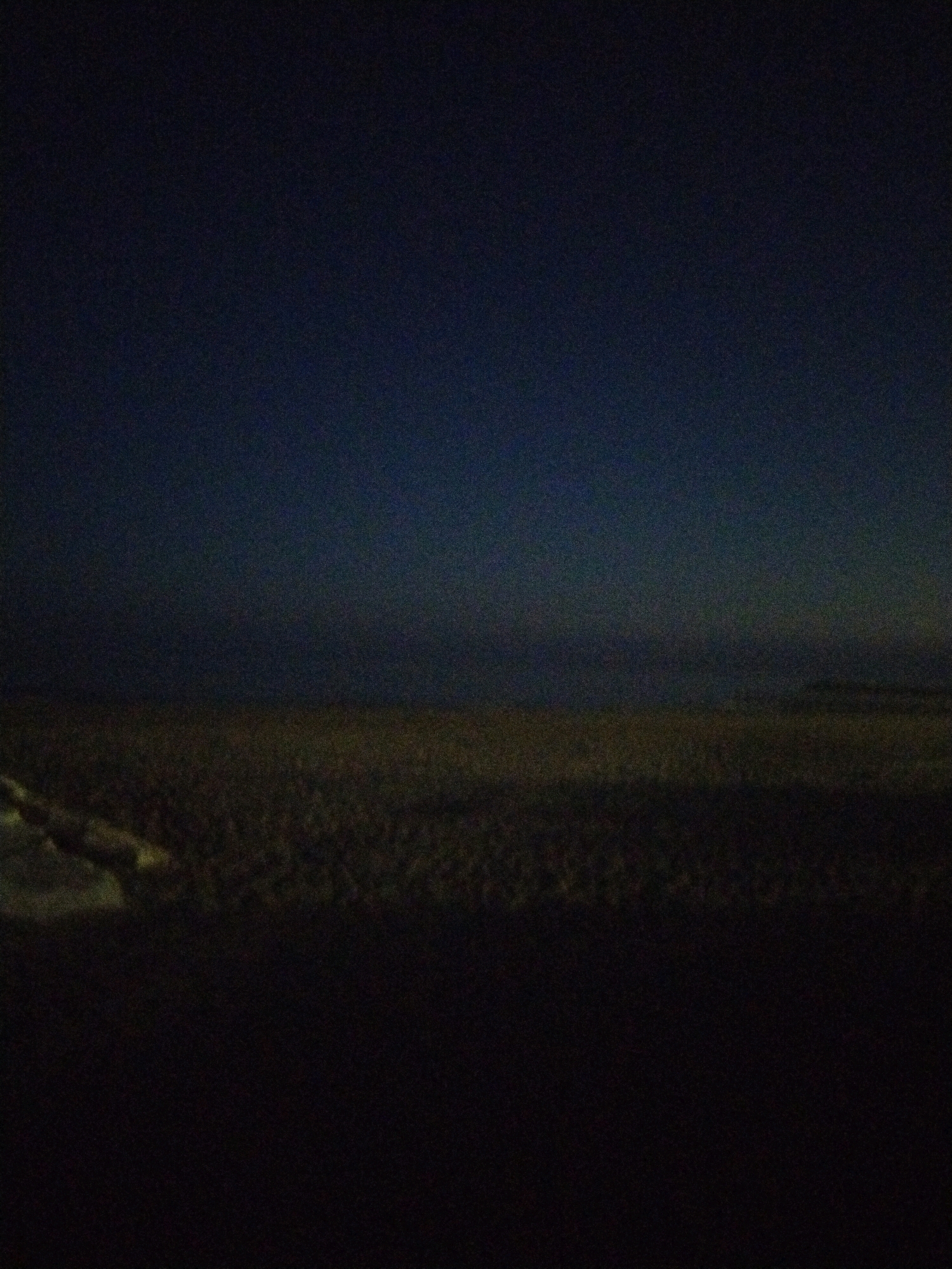 The image depicts a dark, grainy scene at night. A vast, flat expanse of what appears to be a beach or sandy area stretches across the lower half of the frame. The ground is uneven and textured, with some lighter patches suggesting damp sand or exposed stones. In the upper left corner, a faint, illuminated object or area is visible, possibly a road or a path, with a pale, yellowish-white hue. The sky above is a deep, dark blue, transitioning to near black at the top of the image, with no stars or discernible celestial bodies. The overall impression is one of low light conditions, possibly twilight or late night, with limited visibility. There are no people or animals visible in the scene. The location is identified as being in Caorle, Italy, suggesting a coastal environment.